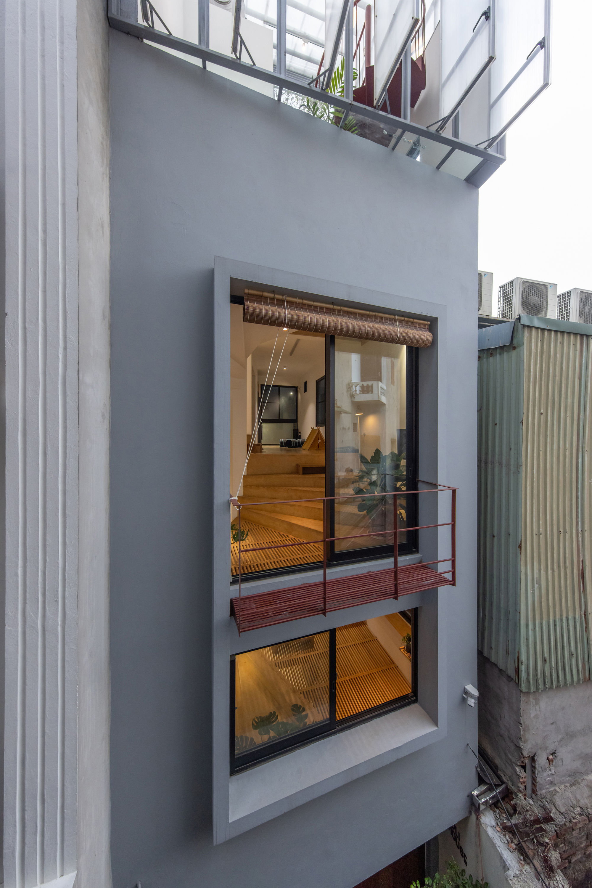 Exterior facade showing stacked glazed openings and a red metal balcony between neighbouring walls at dusk