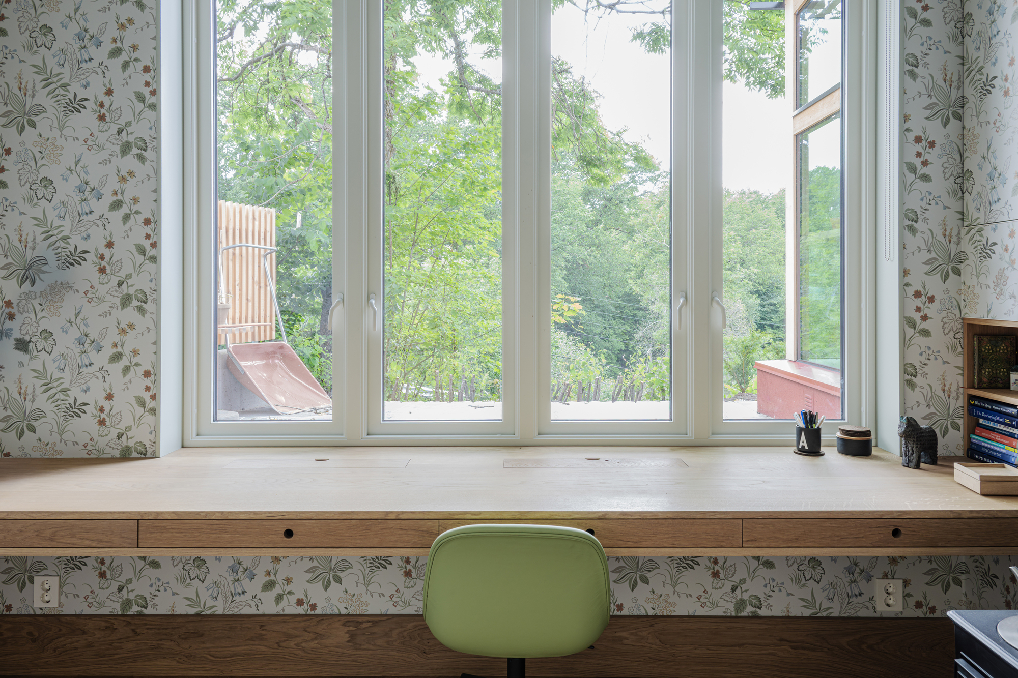 Built-in timber desk below a wide window bay with floral wallpaper and green chair