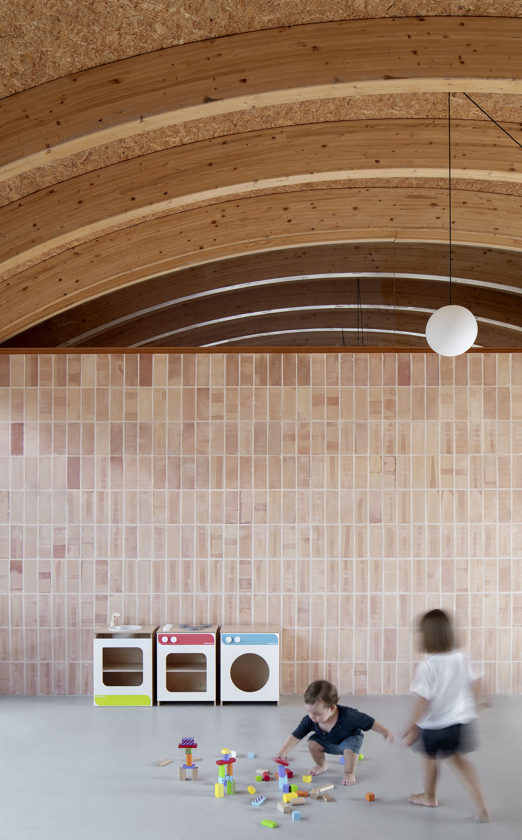 Pink tiled wall with children playing on the polished concrete floor beneath arched timber beams