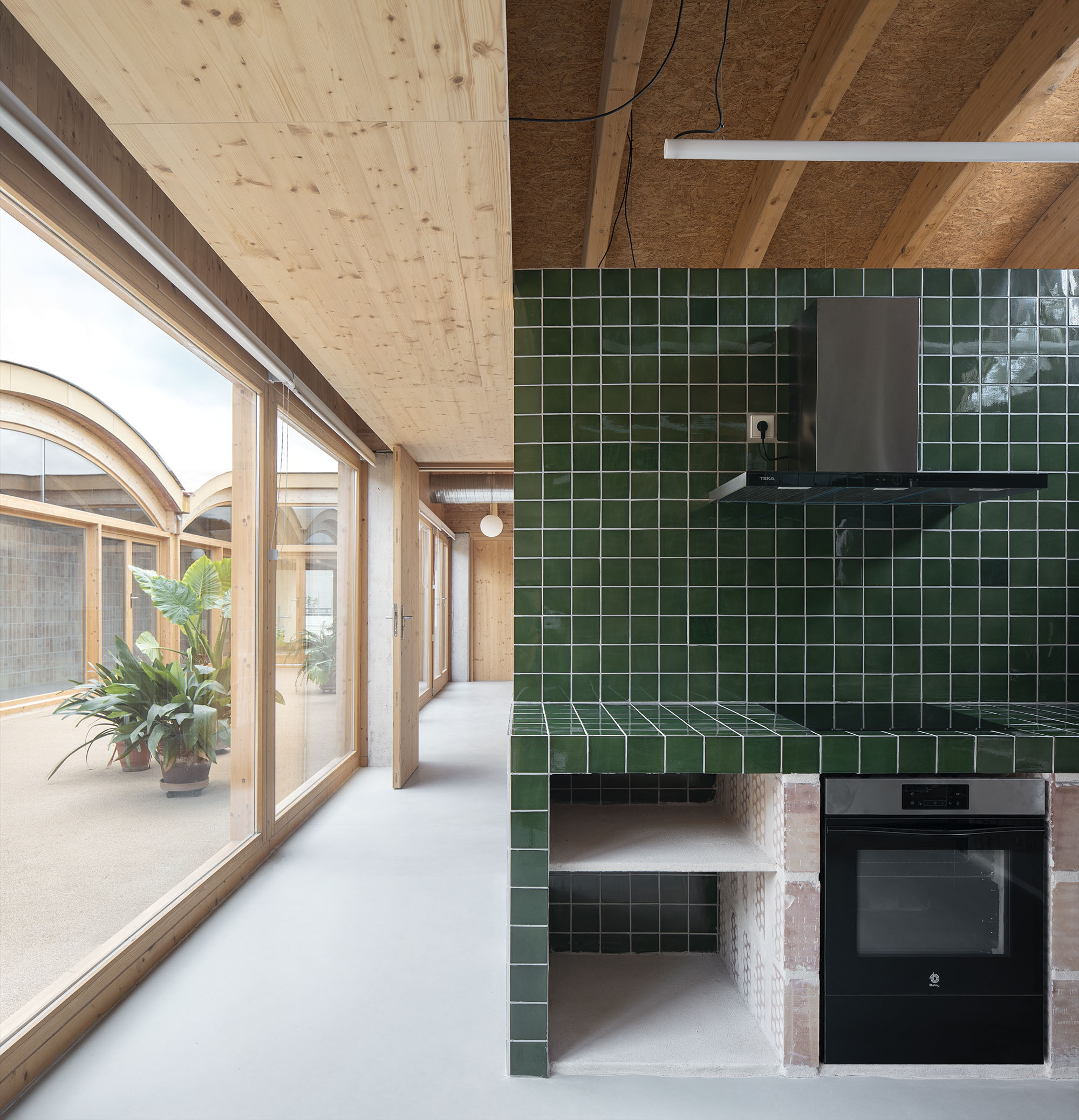 Dark green tiled kitchen volume with built-in oven beneath exposed timber ceiling and arched windows