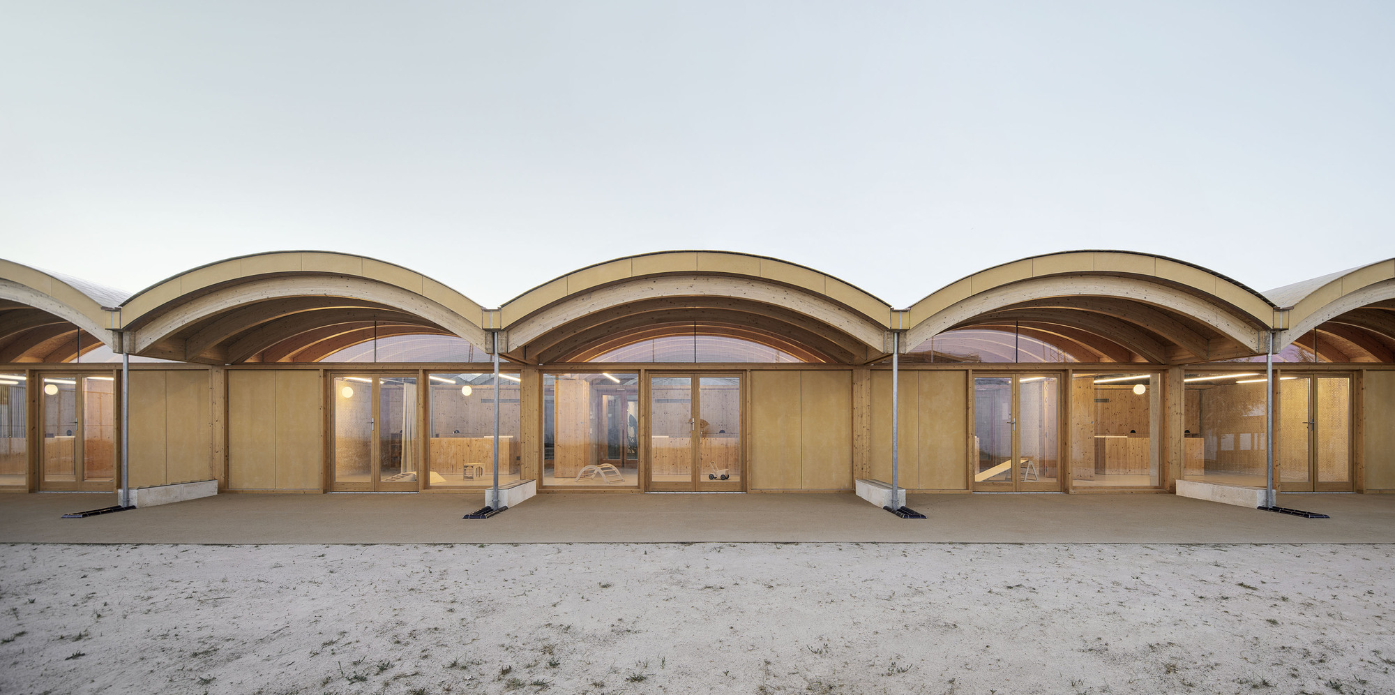 Covered colonnade with repeated arched timber vaults and glazed timber doors opening onto a sandy courtyard