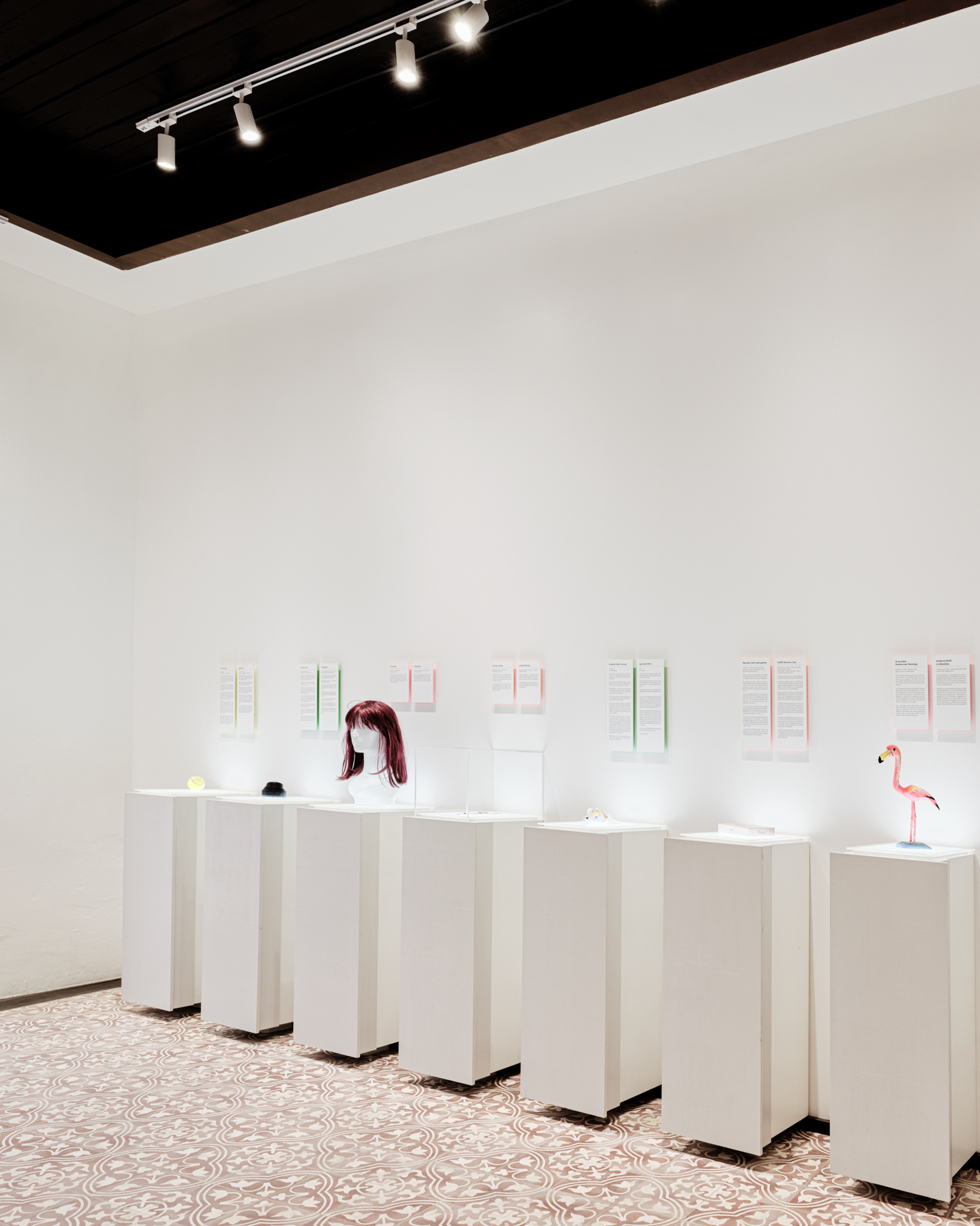 Gallery wall with white pedestals displaying small objects beneath track lighting and patterned floor tiles