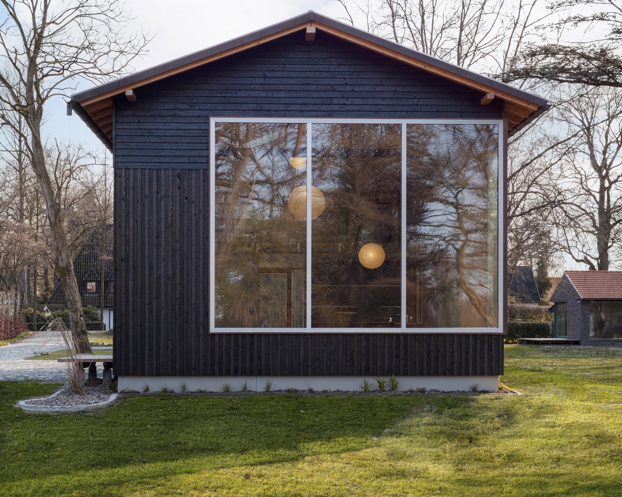 Charred timber gable facade with large tri-panel window reflecting bare trees in late afternoon light