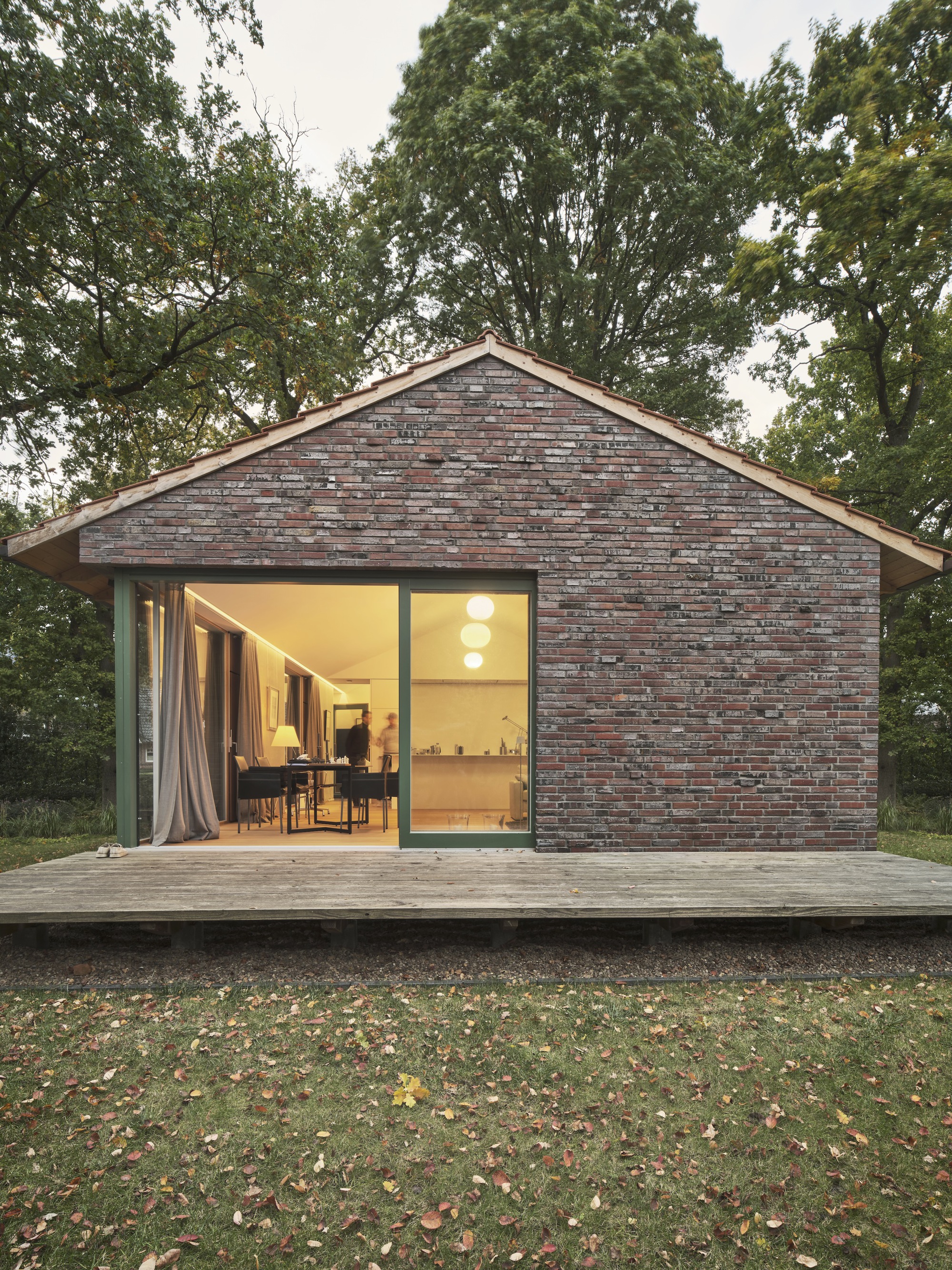 Reclaimed brick gable facade with full-height glazed opening onto timber deck surrounded by fallen autumn leaves