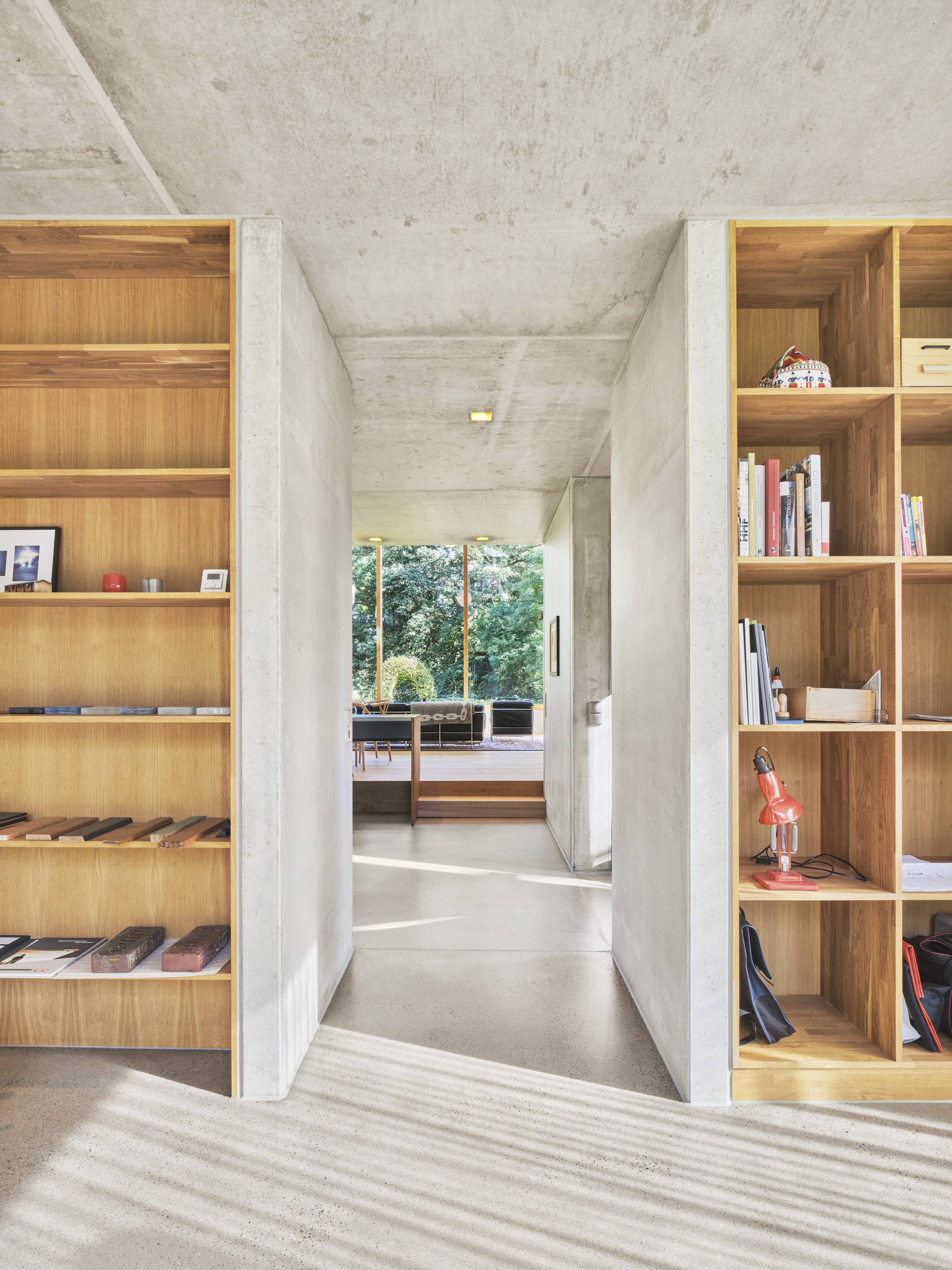Interior hallway with exposed concrete ceiling flanked by built-in timber bookshelves and garden view beyond