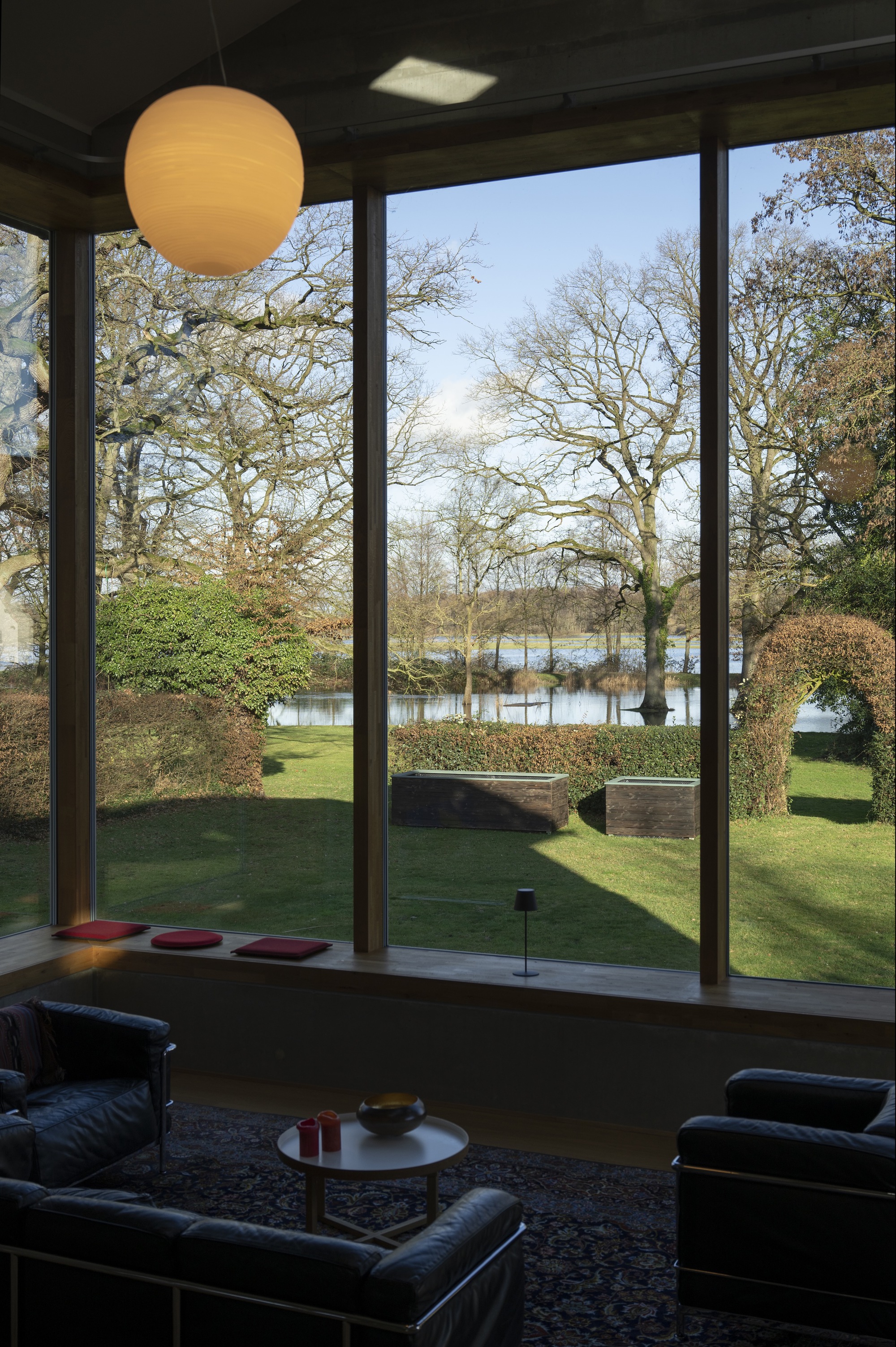 Floor-to-ceiling window framing a view of bare winter trees and a lake beyond a lawn
