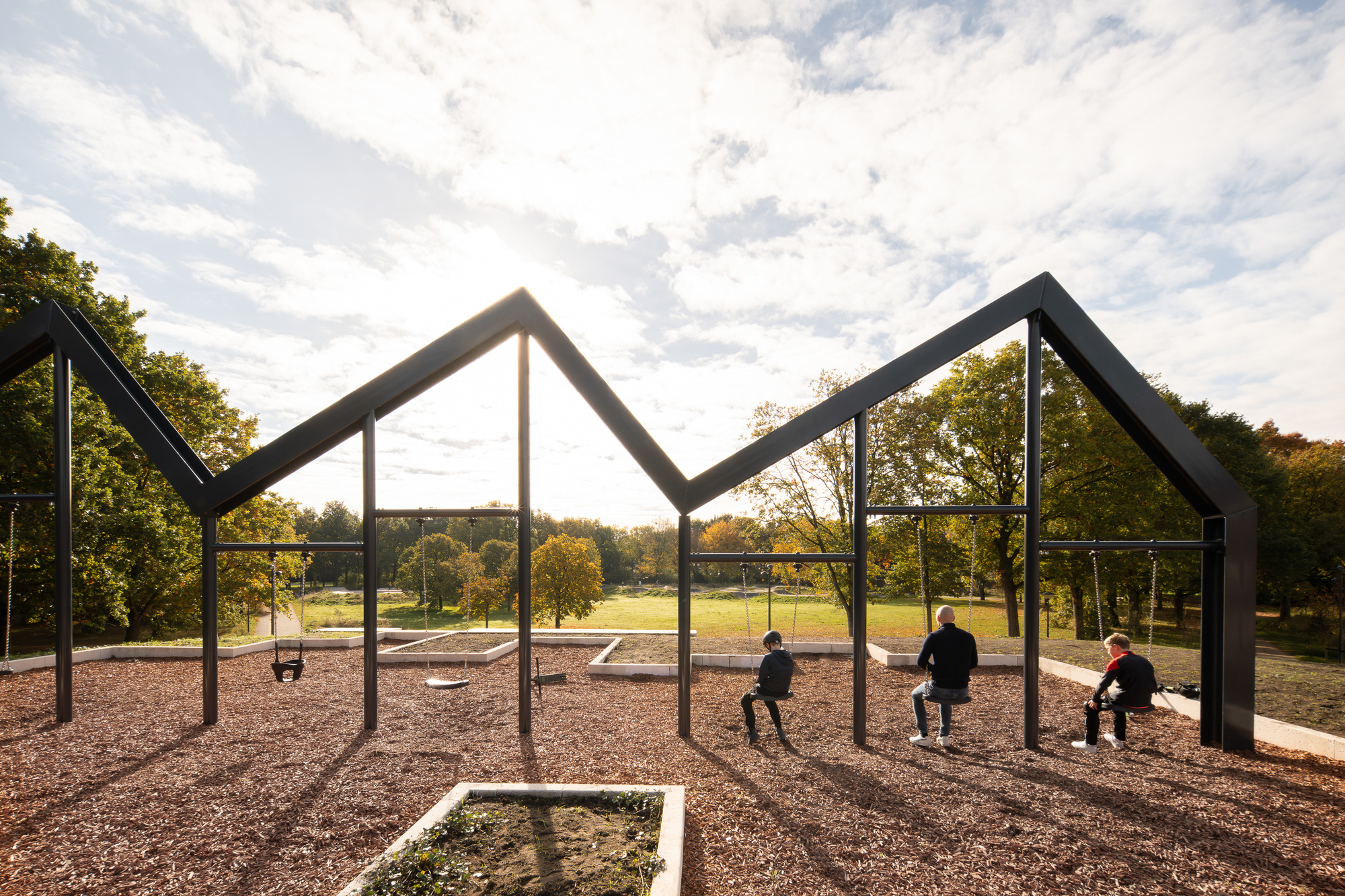 Steel frame house-shaped pavilion with three children seated on swings under a partly cloudy sky