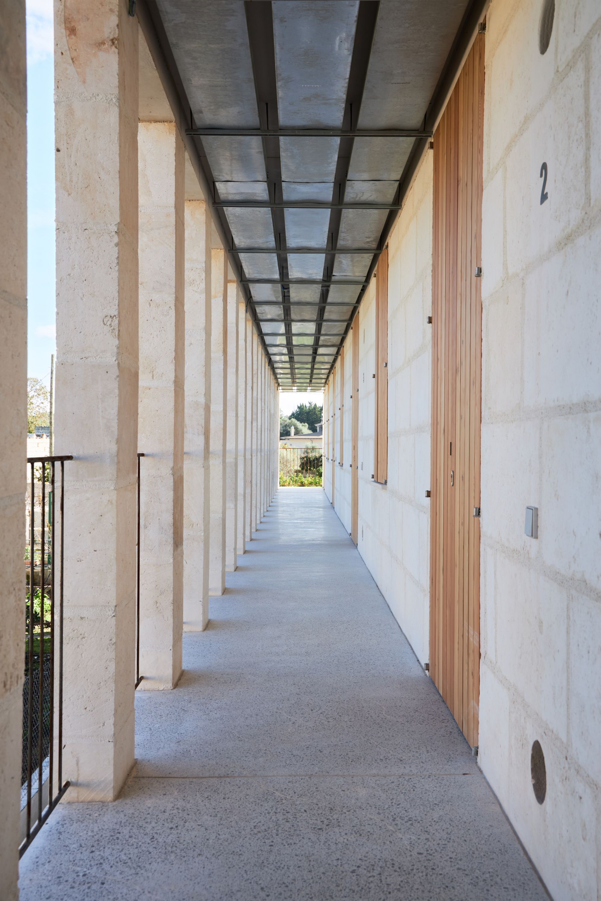 Long exterior corridor with stone columns, steel and glass ceiling panels, and timber doorways along the wall