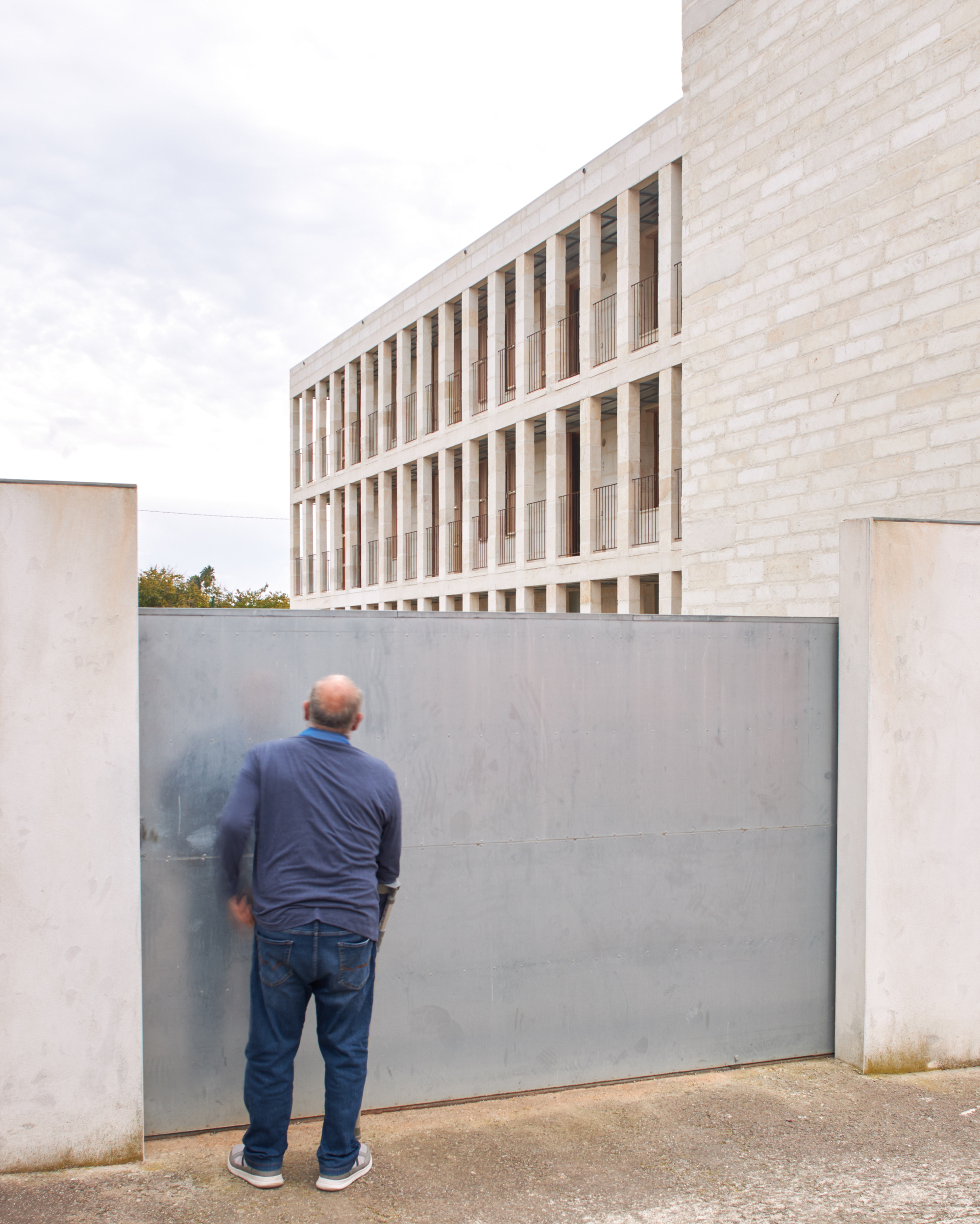 Figure standing at a steel gate below the gridded stone facade in soft light