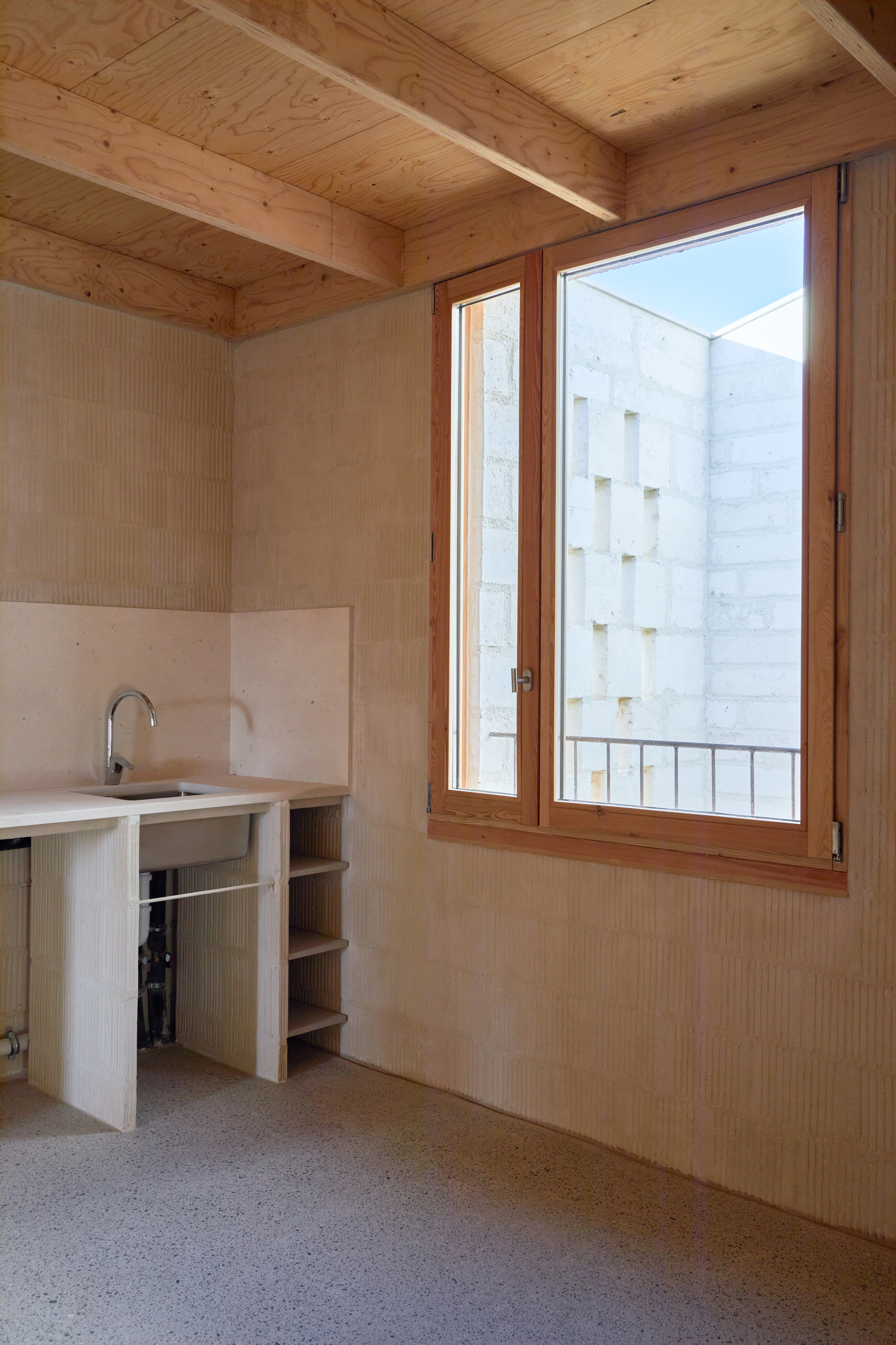 Small kitchen counter with open shelving beneath timber-framed casement window overlooking a neighboring white facade