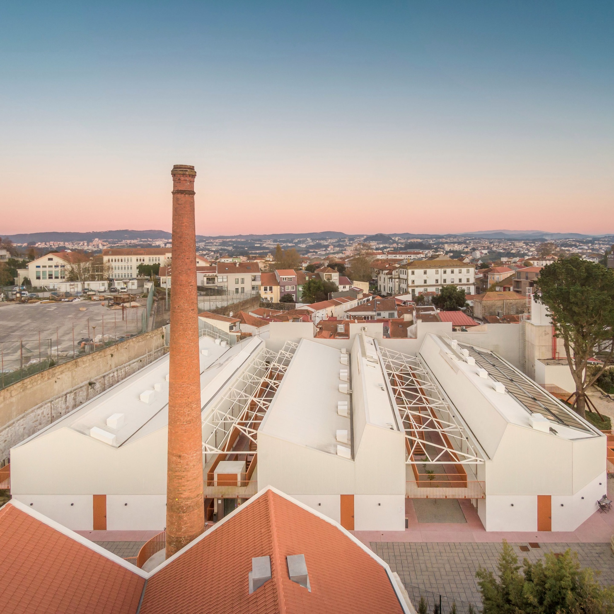 Aerial view of parallel sawtooth roofs with glazed sections and a tall brick chimney at dusk