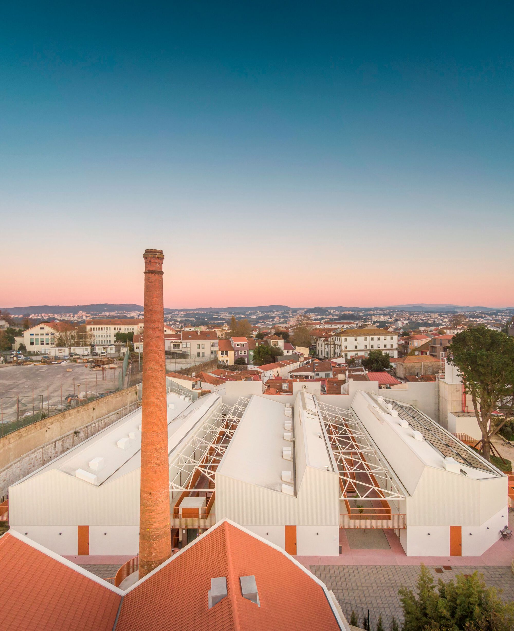 Elevated view showing white sawtooth roofs with exposed steel trusses and a brick chimney at sunset