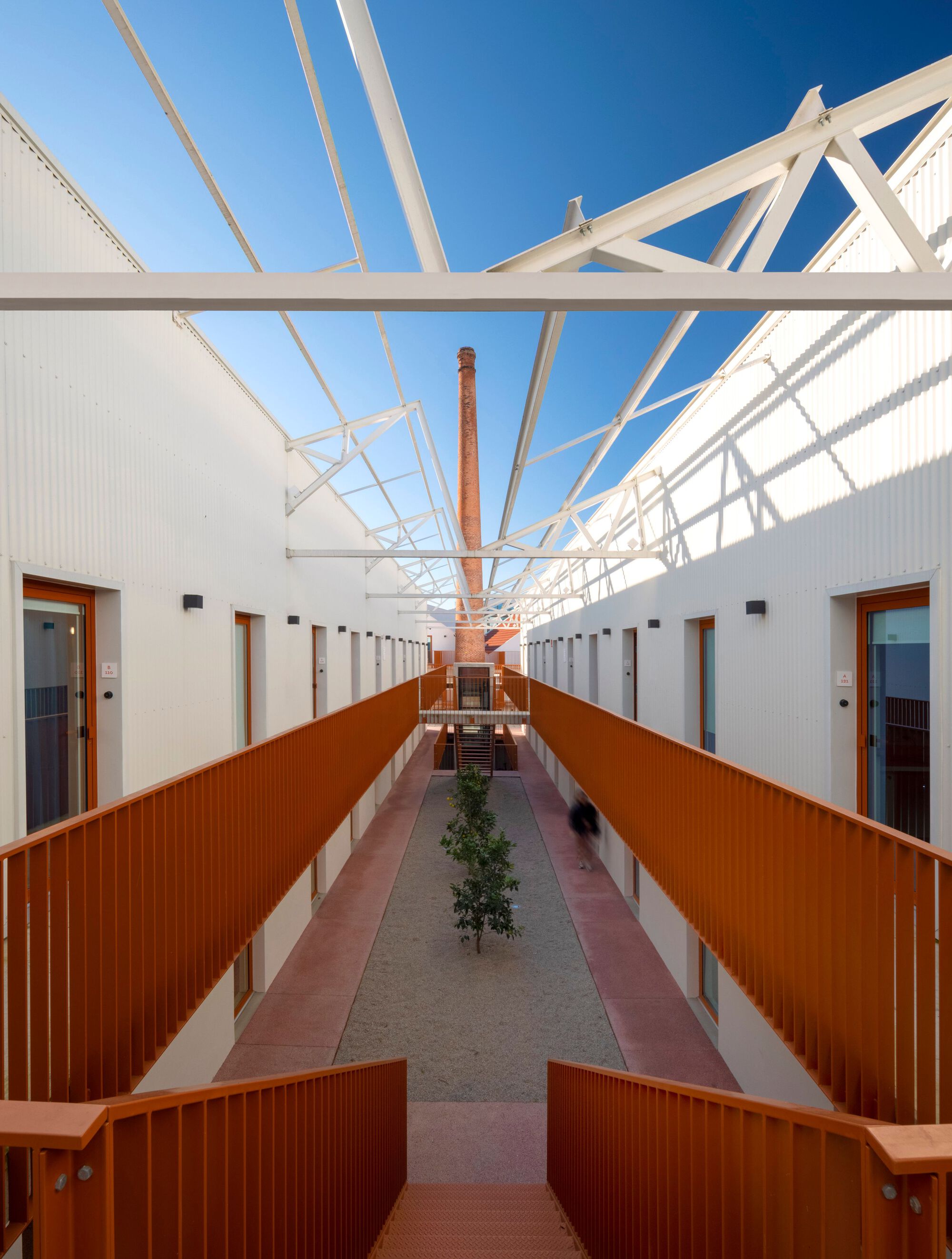 Interior corridor with exposed white timber trusses, terracotta metal railings, and a planted tree