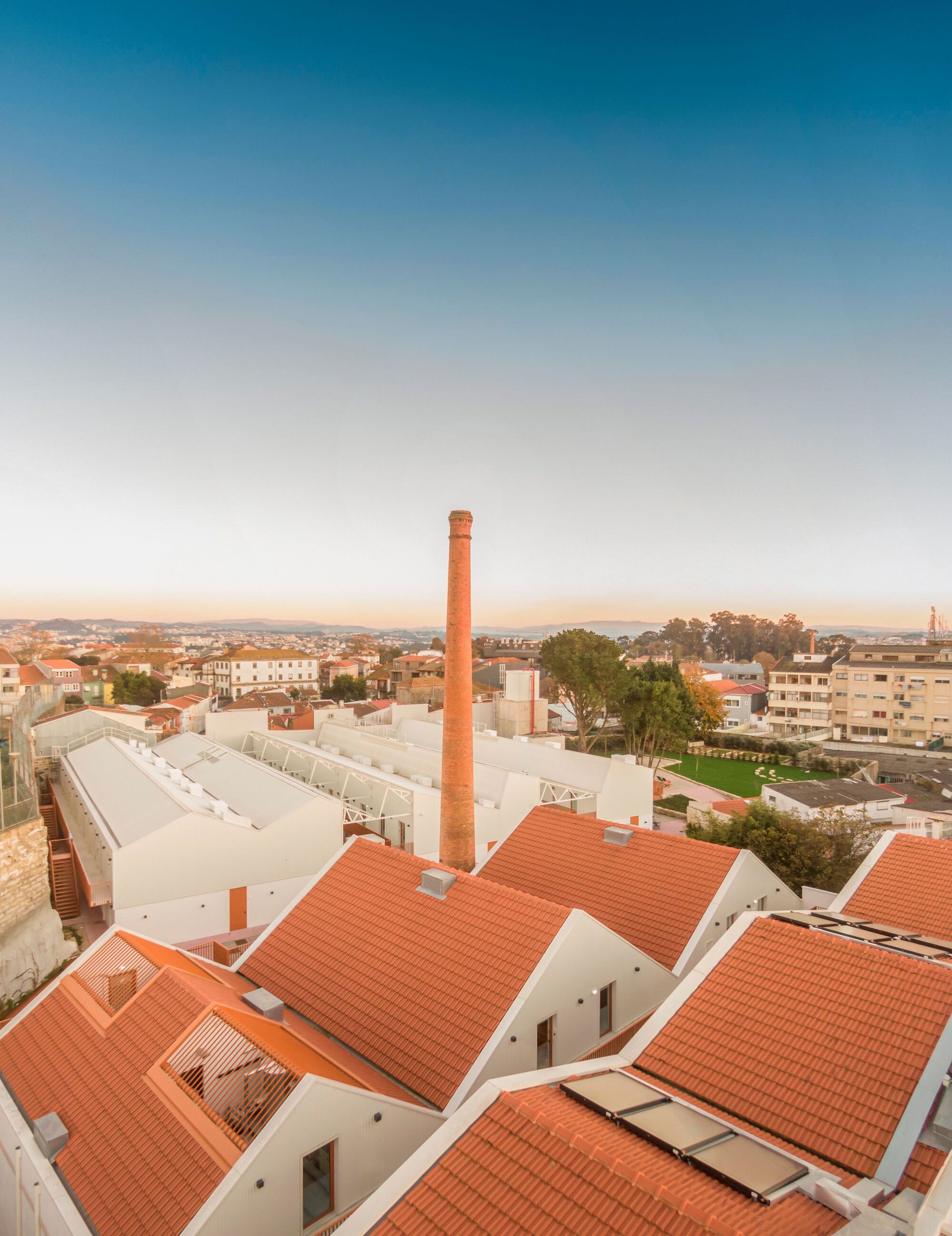 Roofscape with industrial chimney and terracotta-tiled volumes set against low-rise residential surroundings at dusk