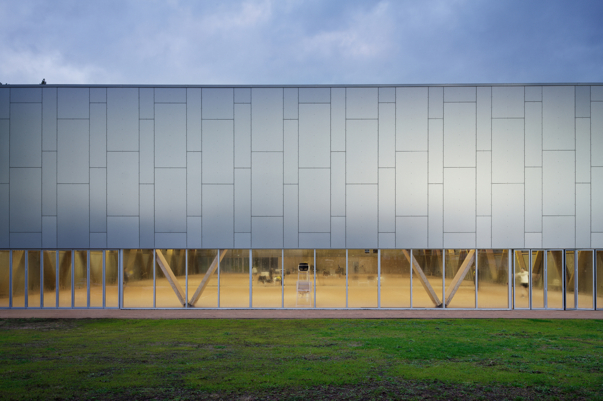 Long facade with pale panel cladding over timber diagonal bracing visible through full-height glazing at dusk