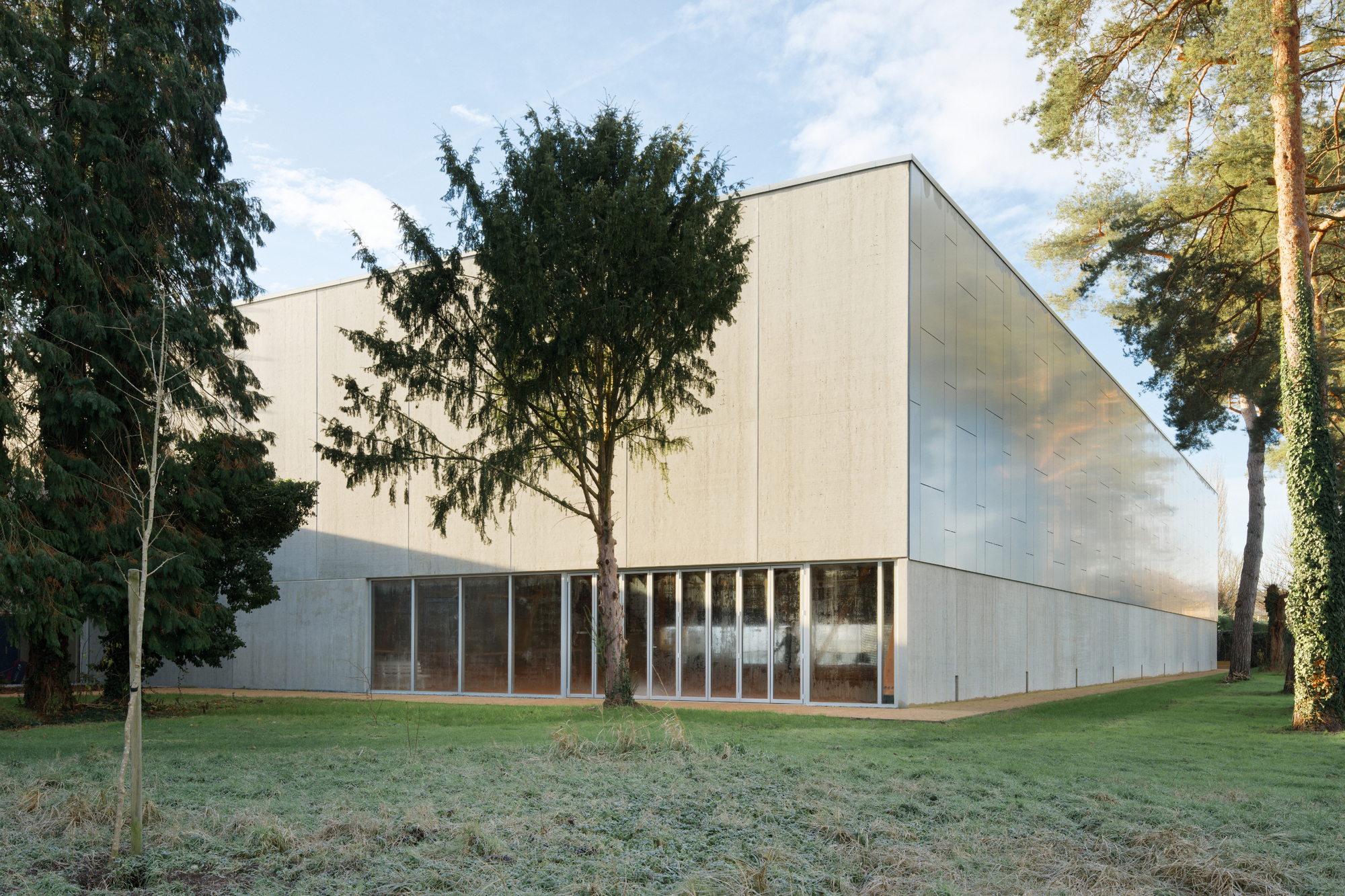 Corner view of the pale metal-clad hall beside mature conifers on frost-covered grass at dawn