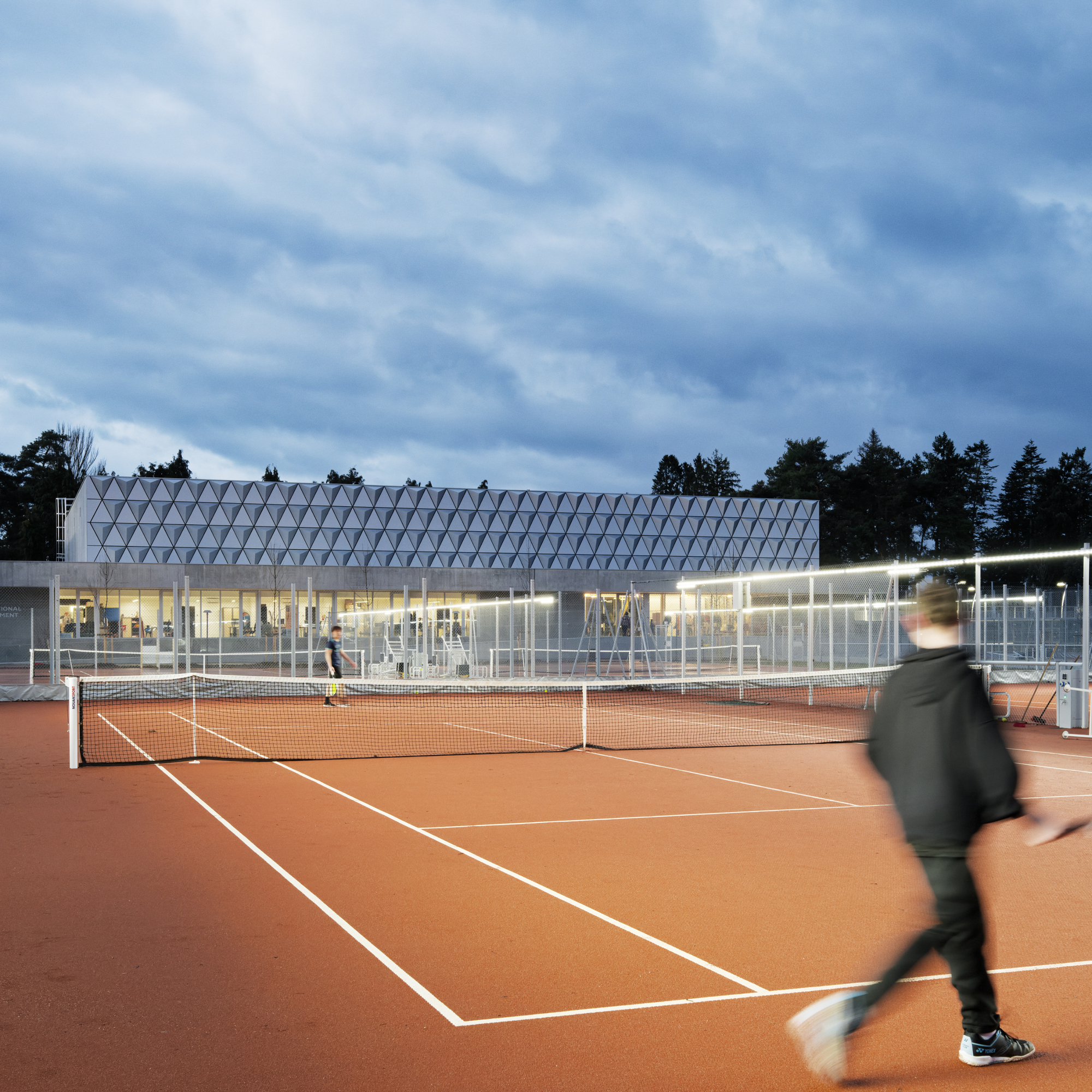 View of the diamond-patterned roof above the tennis pavilion with players on clay courts at dusk