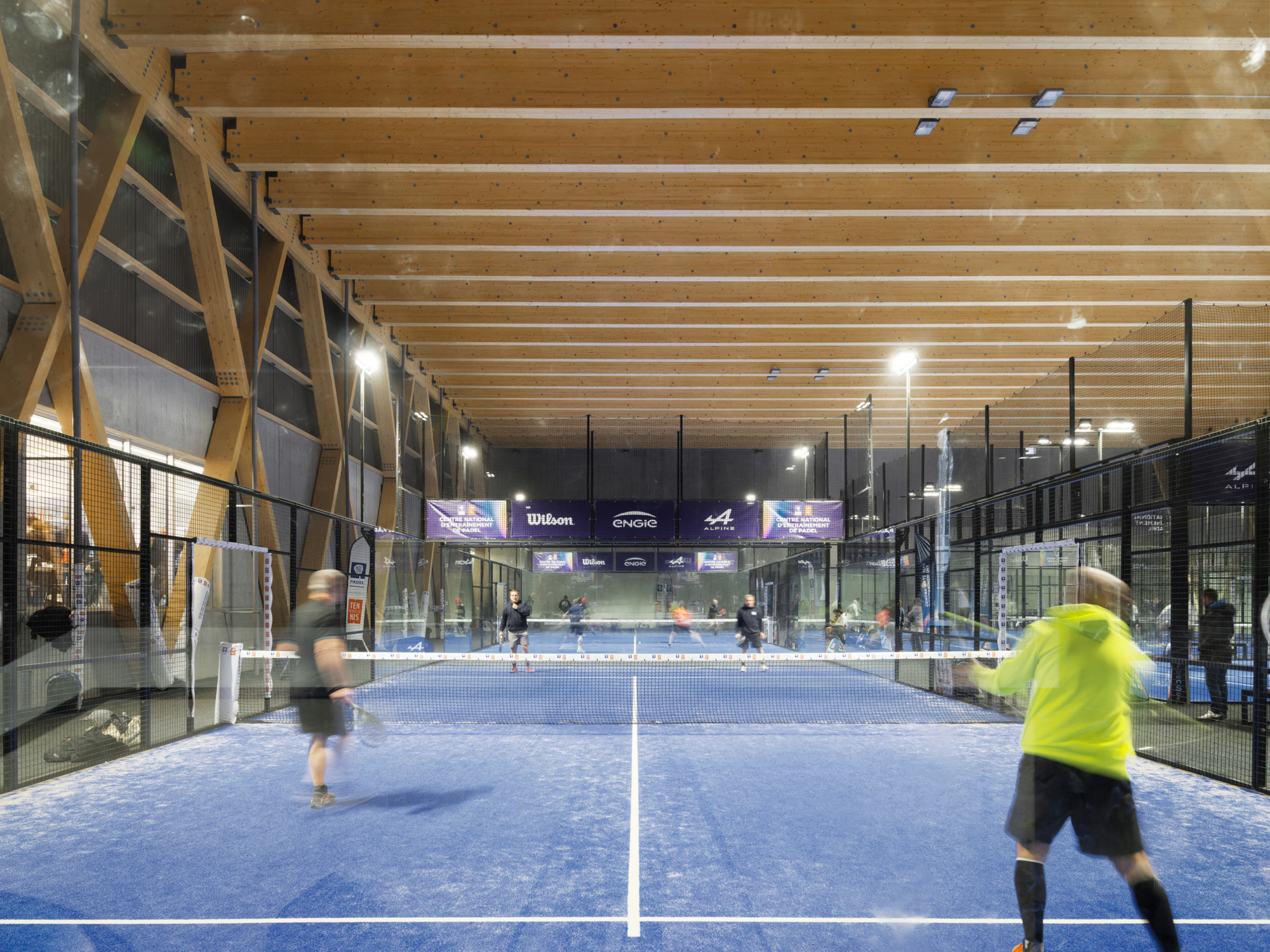 Interior padel court with timber beam ceiling and players under artificial lighting