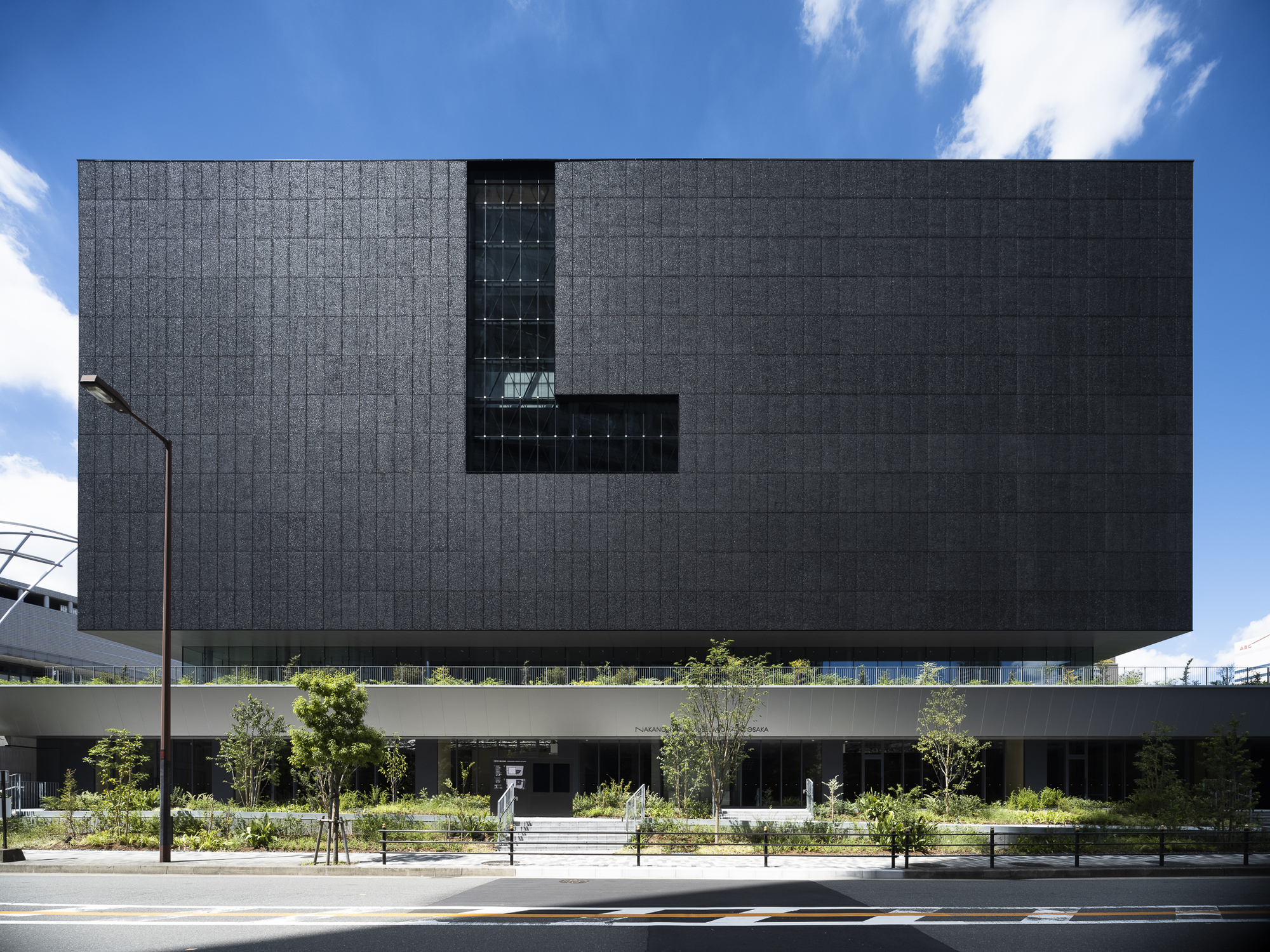 Street view of the dark tiled facade elevated above a landscaped plaza with young trees