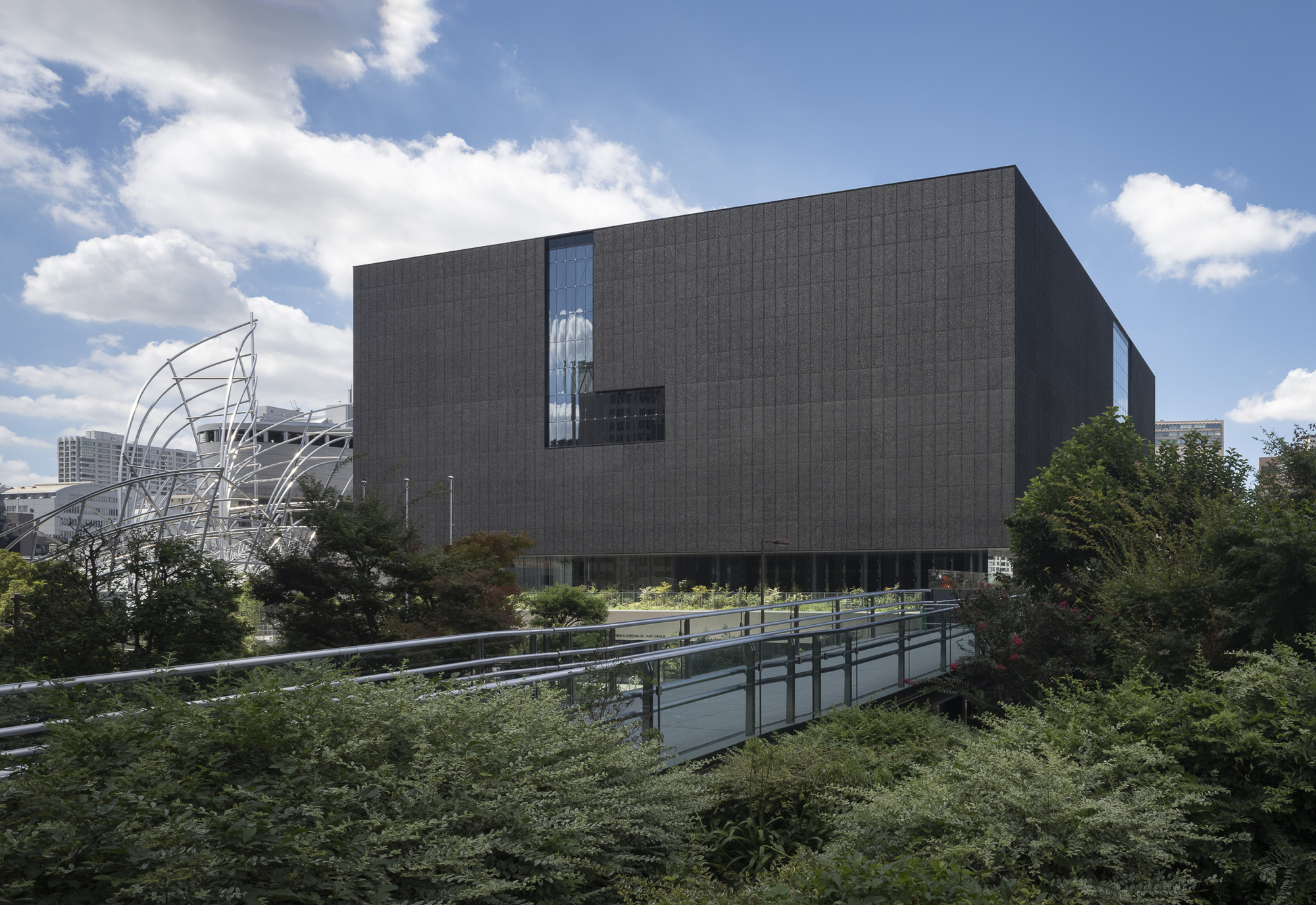 Corner view showing the textured dark facade and elevated volume above a garden with accessible ramp