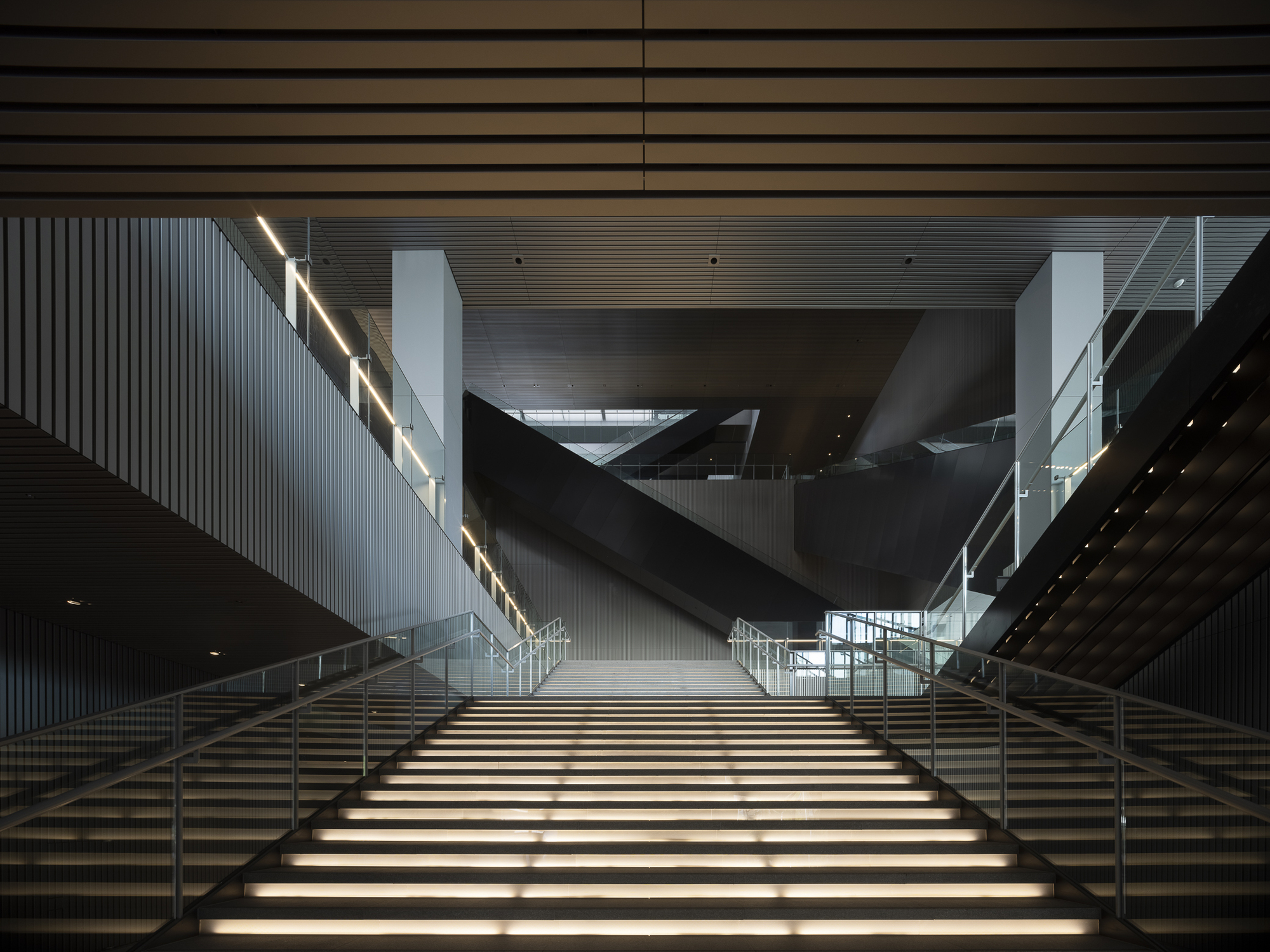 Wide staircase with integrated lighting rising toward angular volumes suspended in the atrium