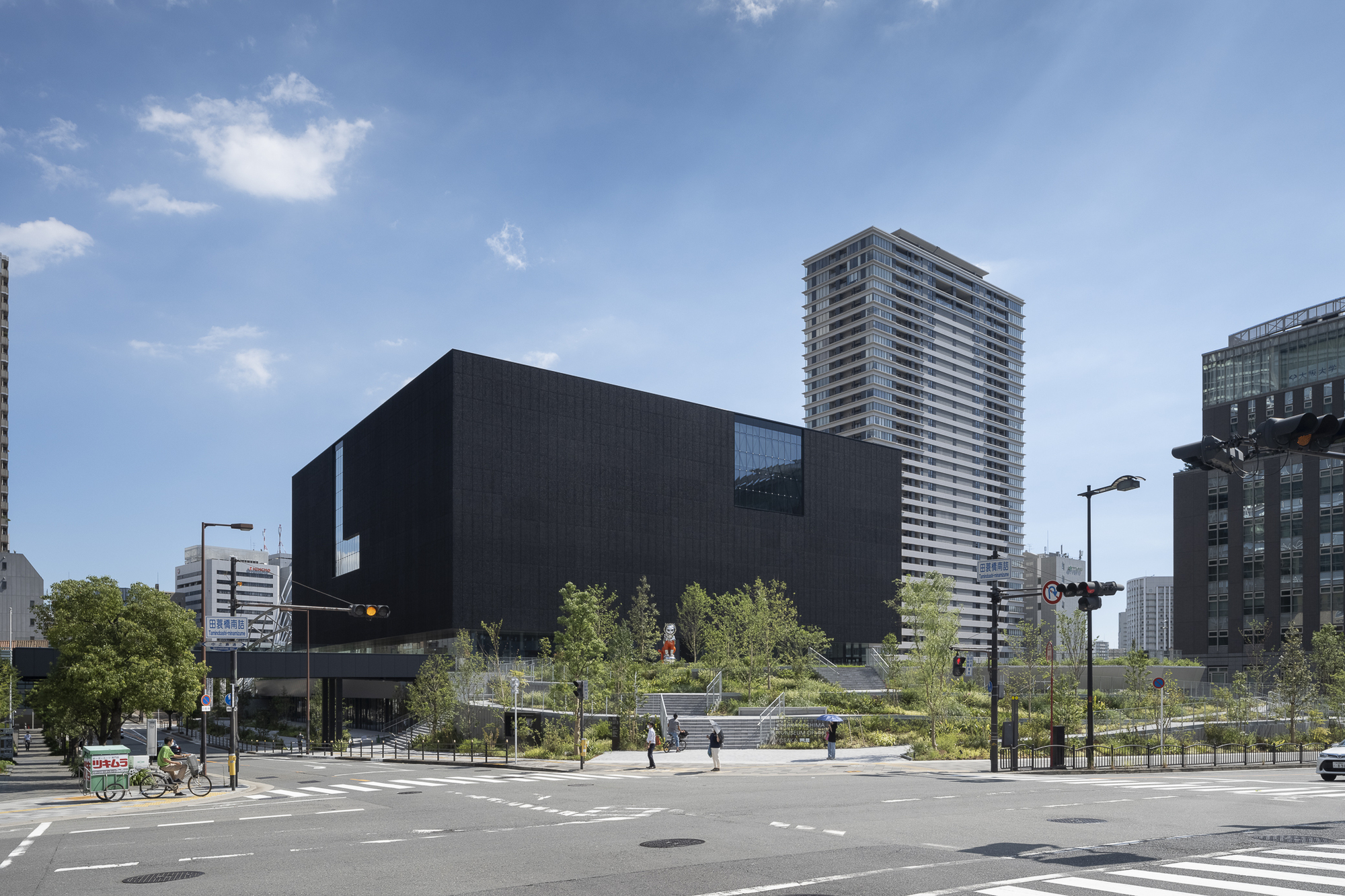 Street corner view of the black volume elevated above a plaza with cyclists and pedestrians