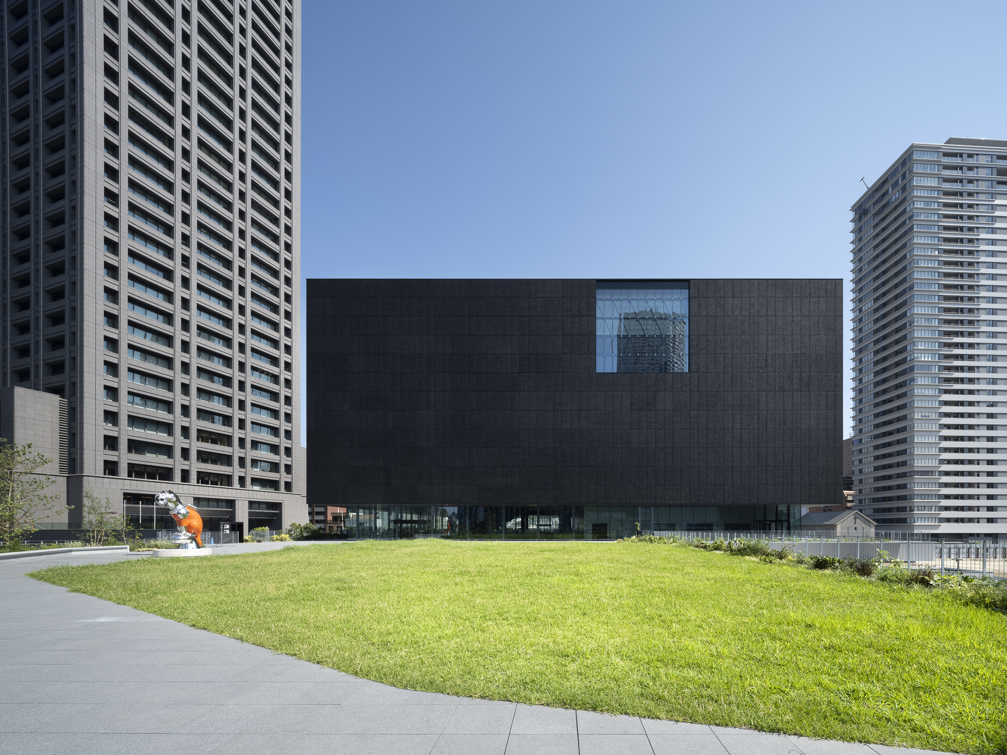 Dark textured facade with single recessed window facing a lawn and residential towers beyond
