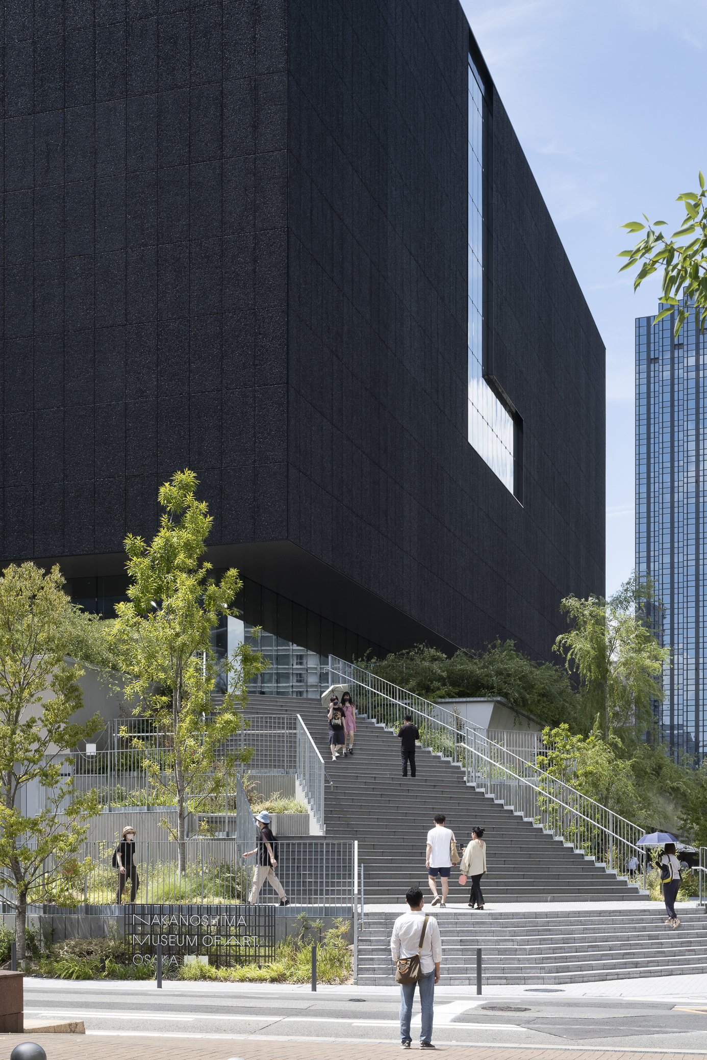 Wide staircase with visitors ascending beneath the cantilevered dark mass and surrounding greenery