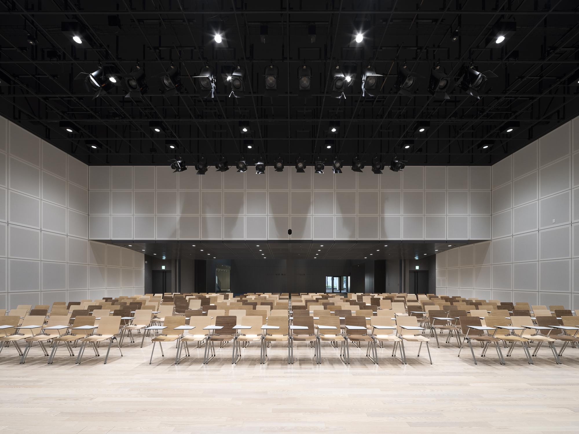 Performance hall with exposed rigging, white panel walls and wooden seating on pale flooring