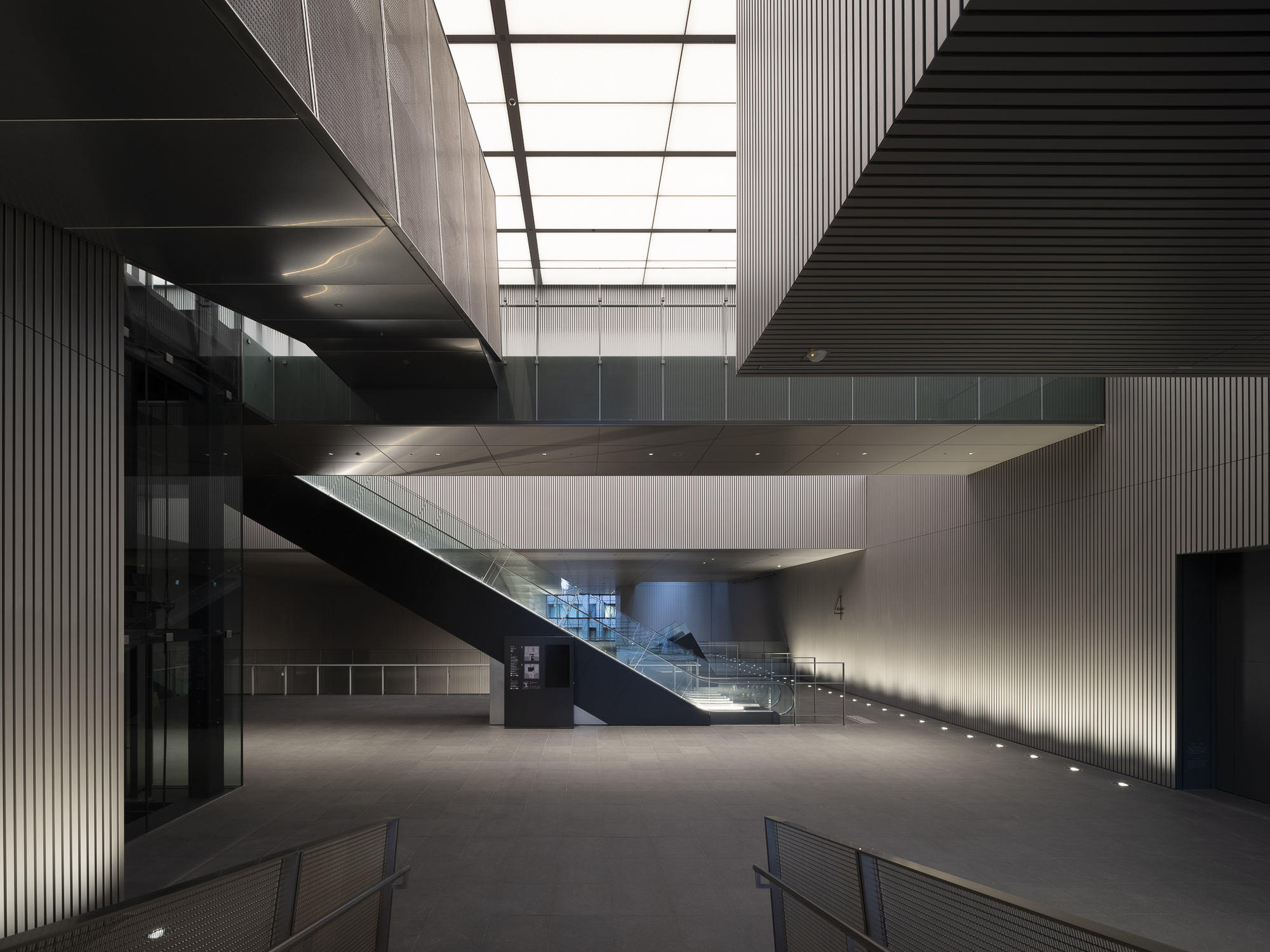 Ground floor lobby with cantilevered stair platform hovering above under a gridded skylight