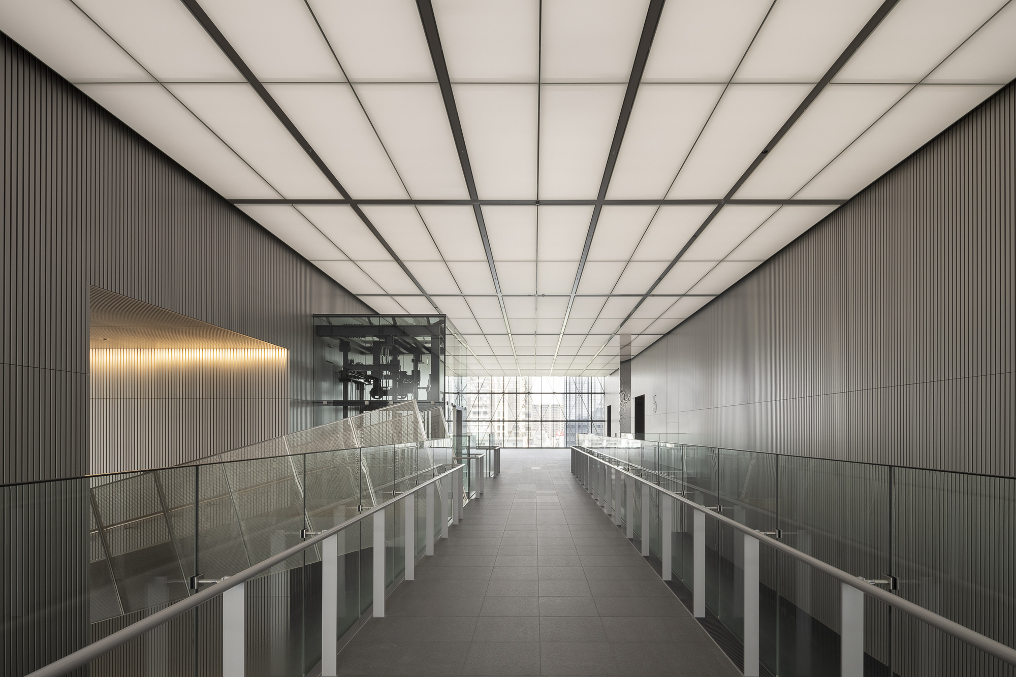 Interior bridge with glass railings under a gridded translucent ceiling framed by ribbed metal walls