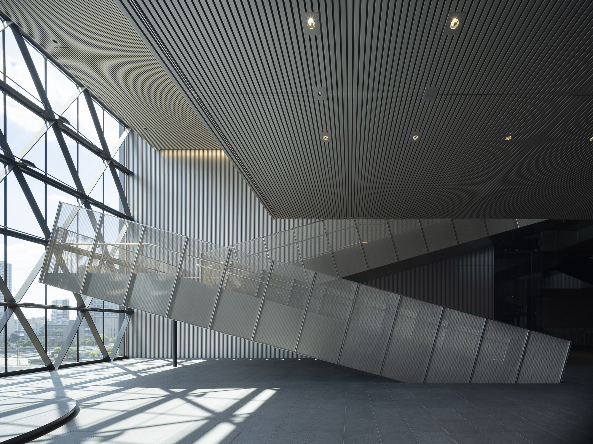 Diagonal steel bracing meets glass and ribbed ceiling panels with sunlight casting sharp shadows across the floor
