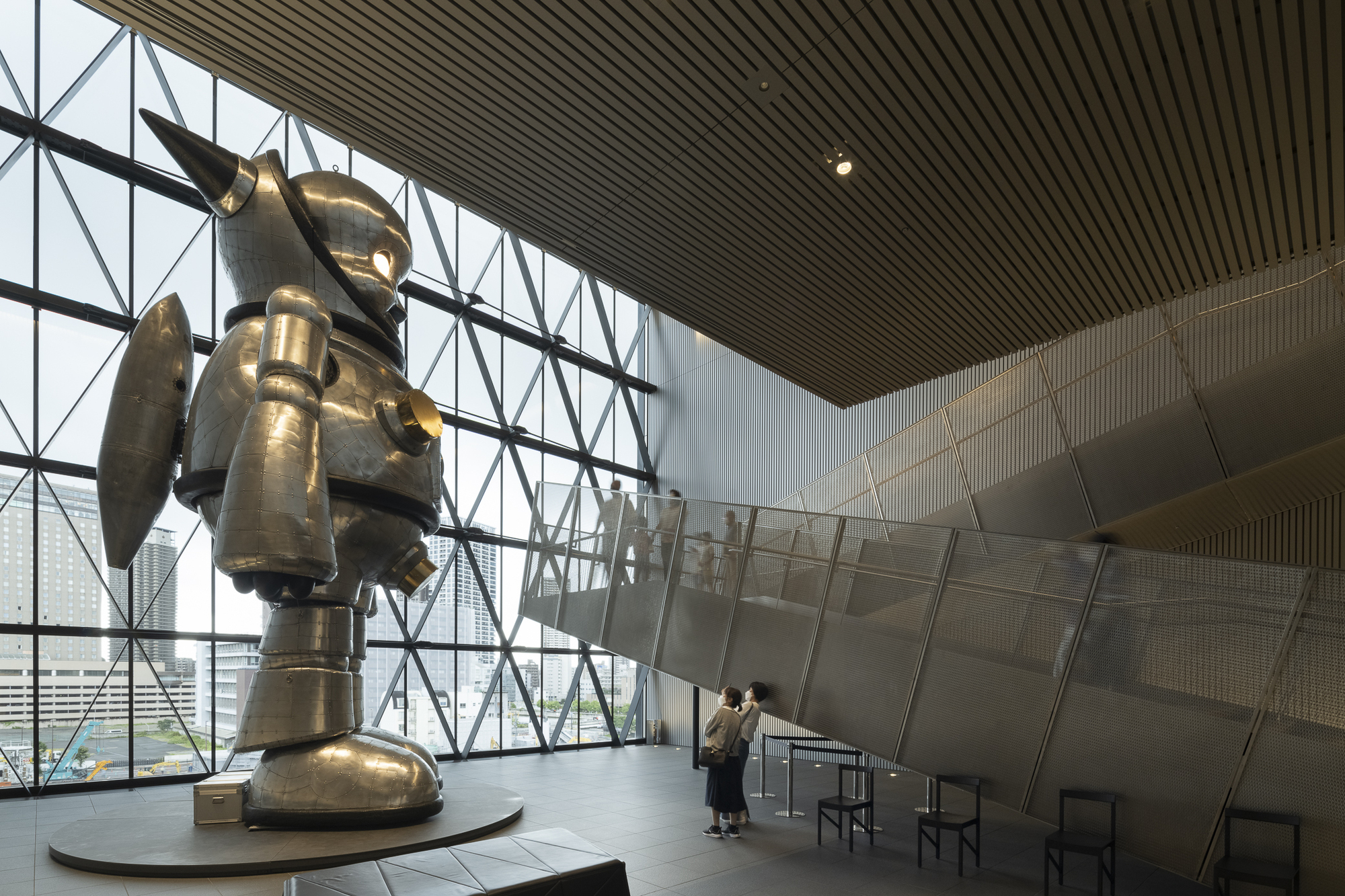 Large bronze sculpture on a circular plinth beneath diagonal steel truss glazing with visitors nearby