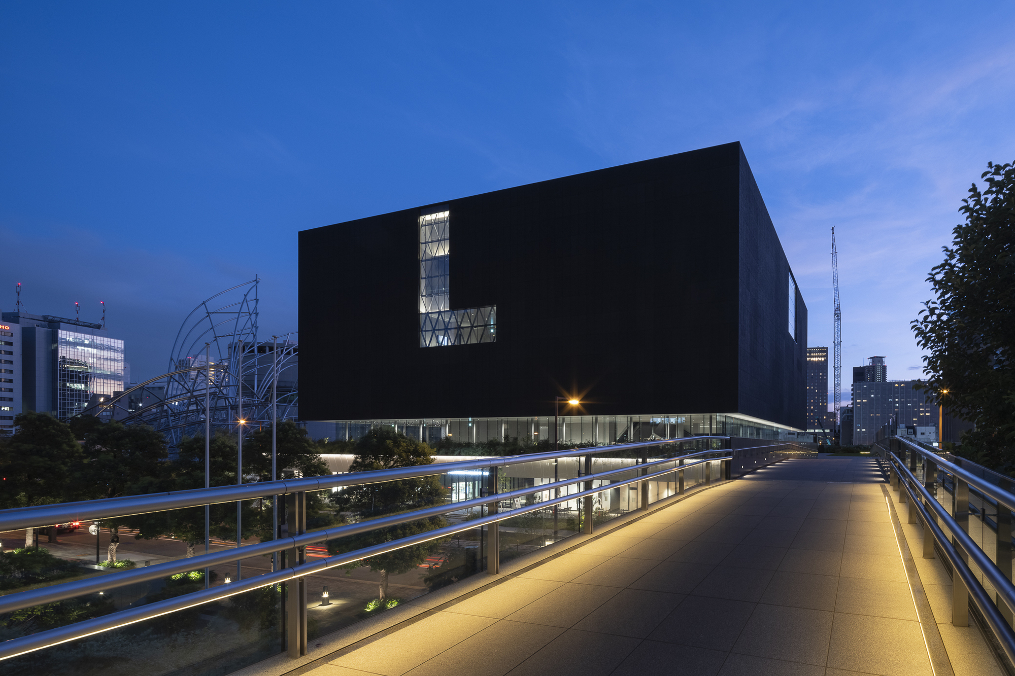 Black cubic volume elevated on slender columns viewed from pedestrian bridge at dusk
