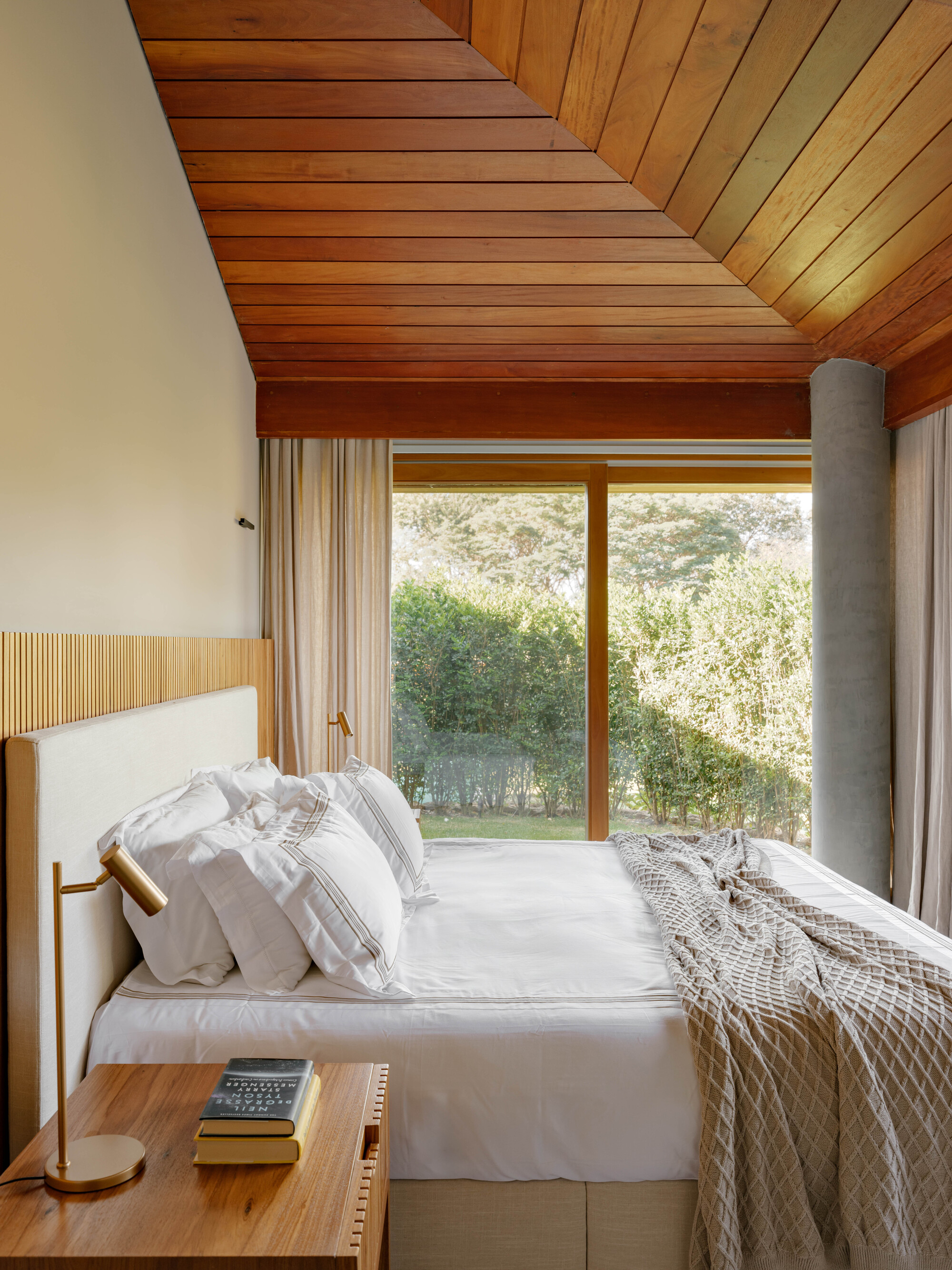 Bedroom with timber slatted ceiling, timber-framed glazing overlooking garden and cylindrical concrete flue column