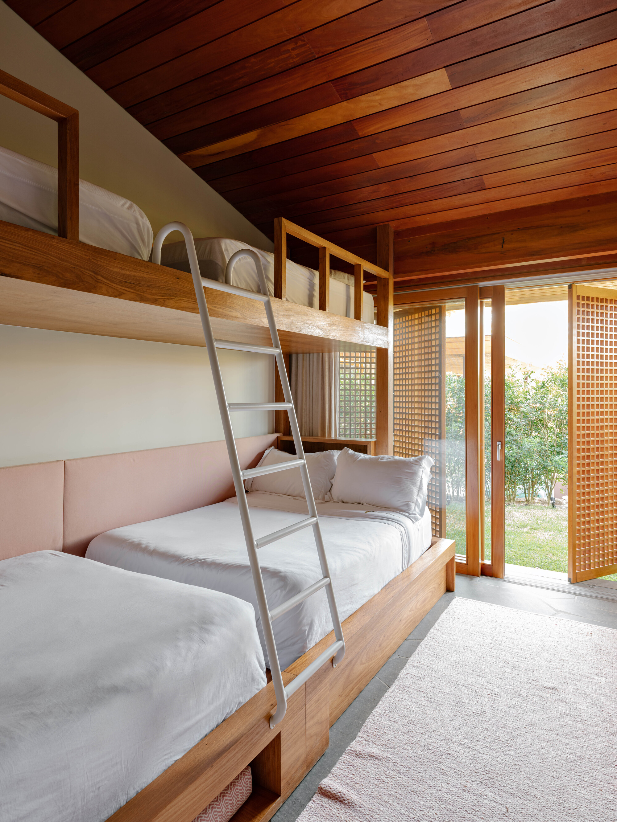 Bunk bedroom with timber-framed beds, metal ladder, sloped timber ceiling and screened sliding doors to garden