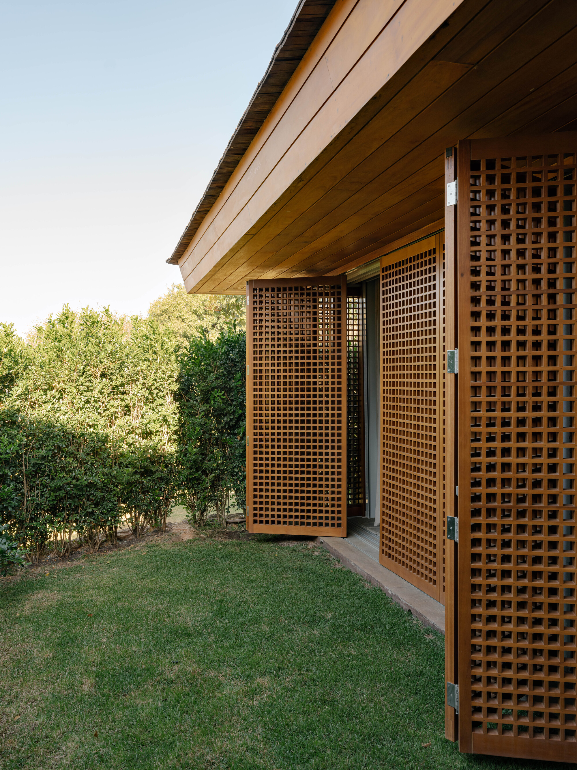 Exterior view of timber lattice screen doors opening onto lawn with timber soffit and dense hedge beyond