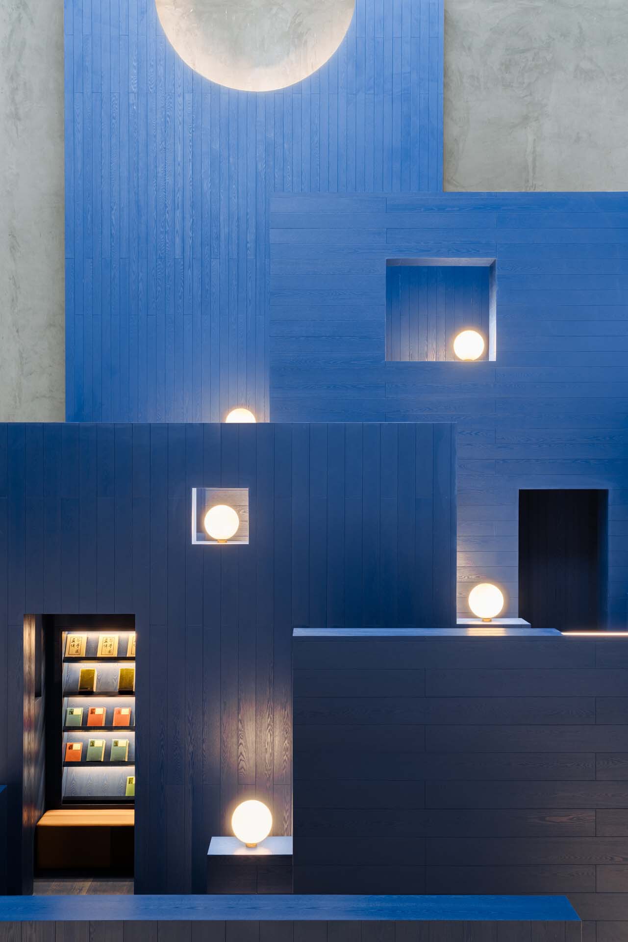 Blue painted timber volumes with niched spherical lamps and a book display visible at dusk