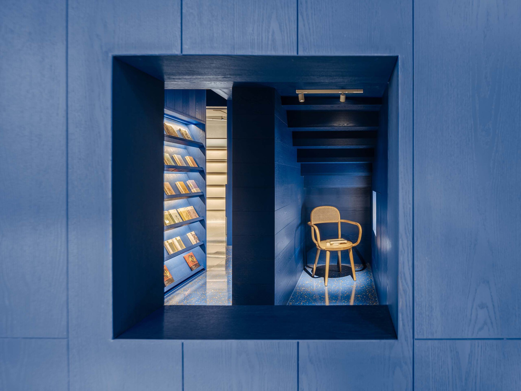 Framed view through blue wood paneling revealing illuminated shelves and a single bentwood chair