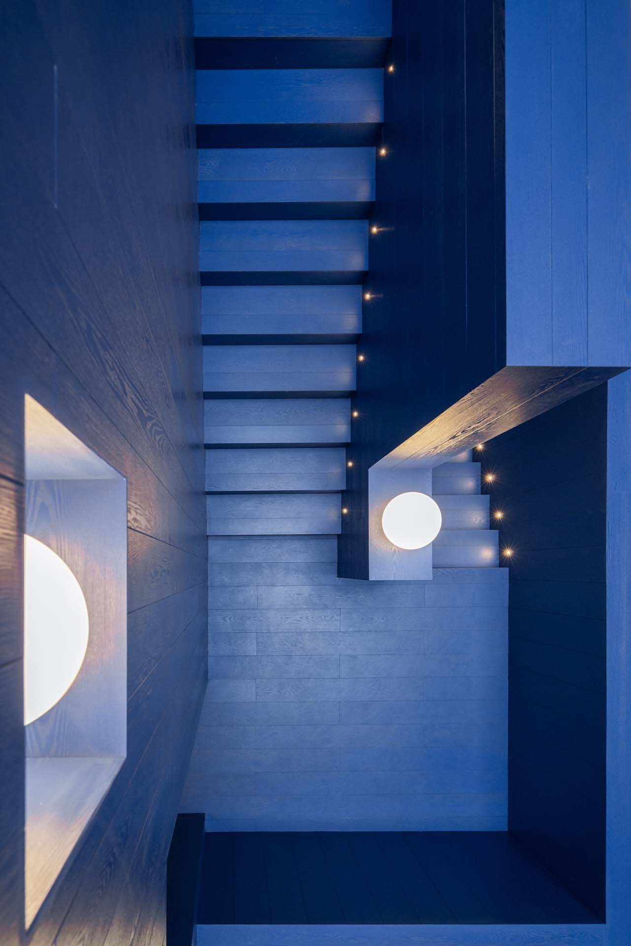 Top-down view of a suspended staircase with illuminated treads and circular ceiling openings at dusk
