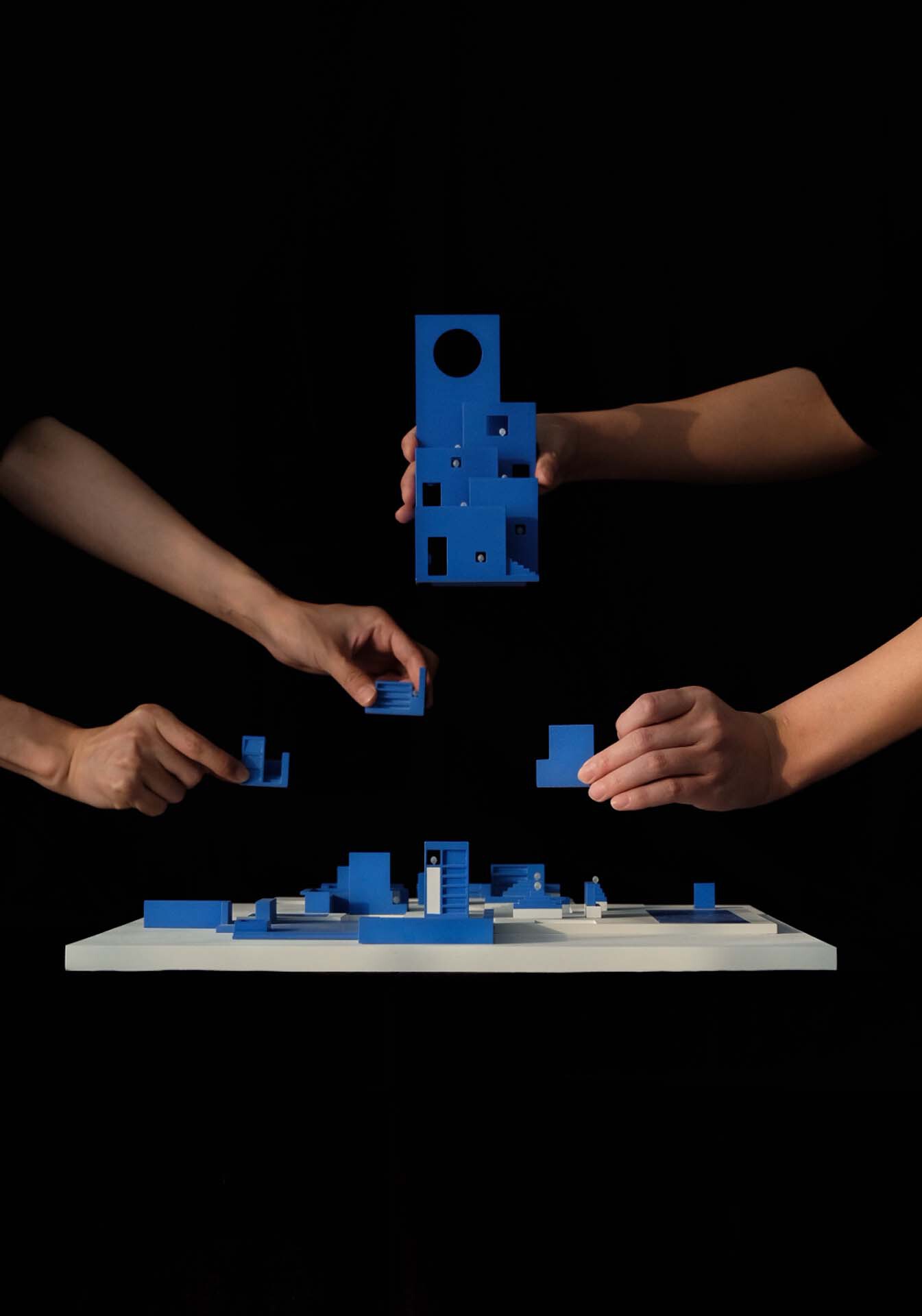 Multiple hands assembling blue modular architectural components above a white base model against black backdrop