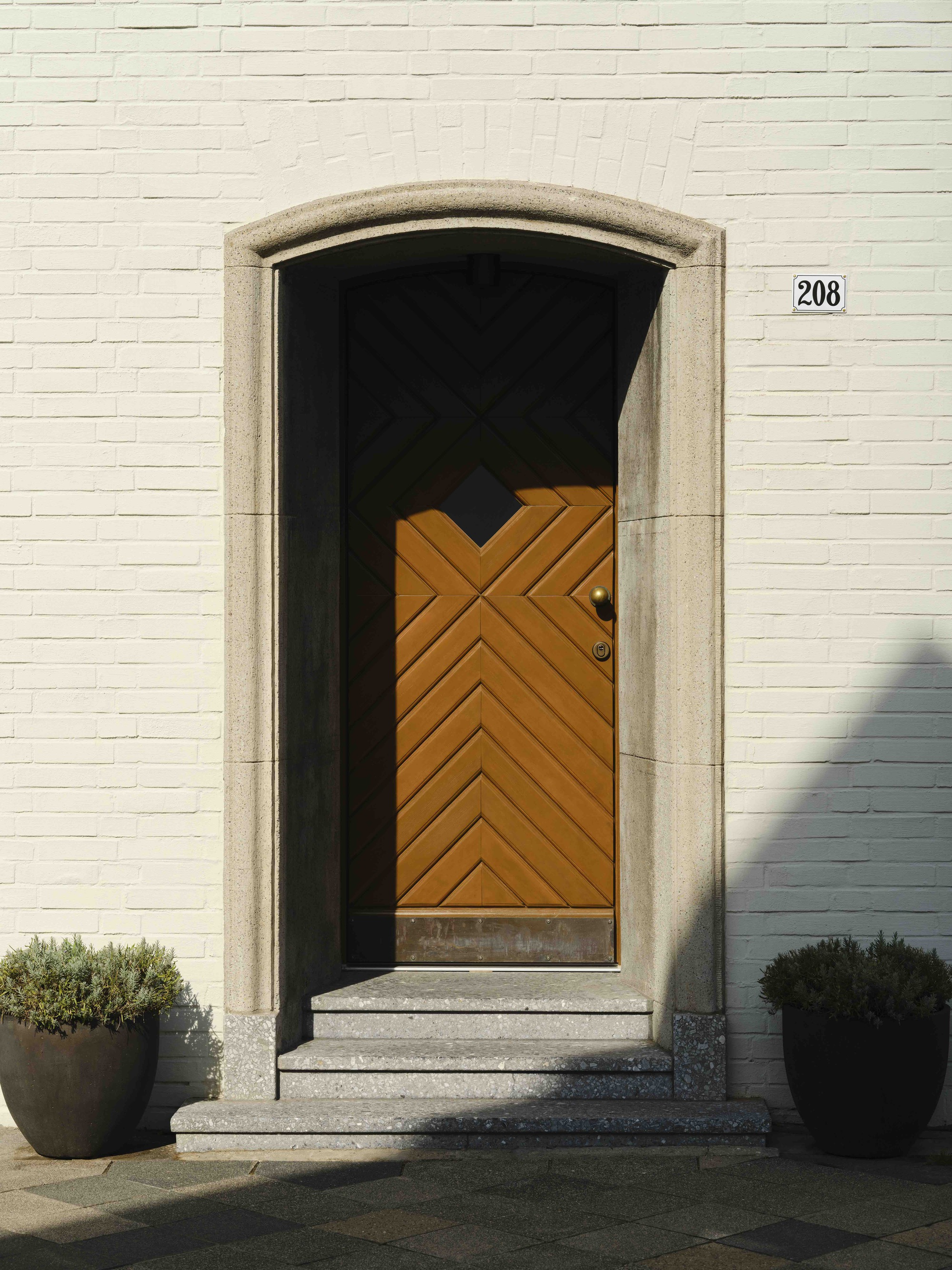Arched doorway with chevron-patterned timber door set in white brick facade flanked by planters