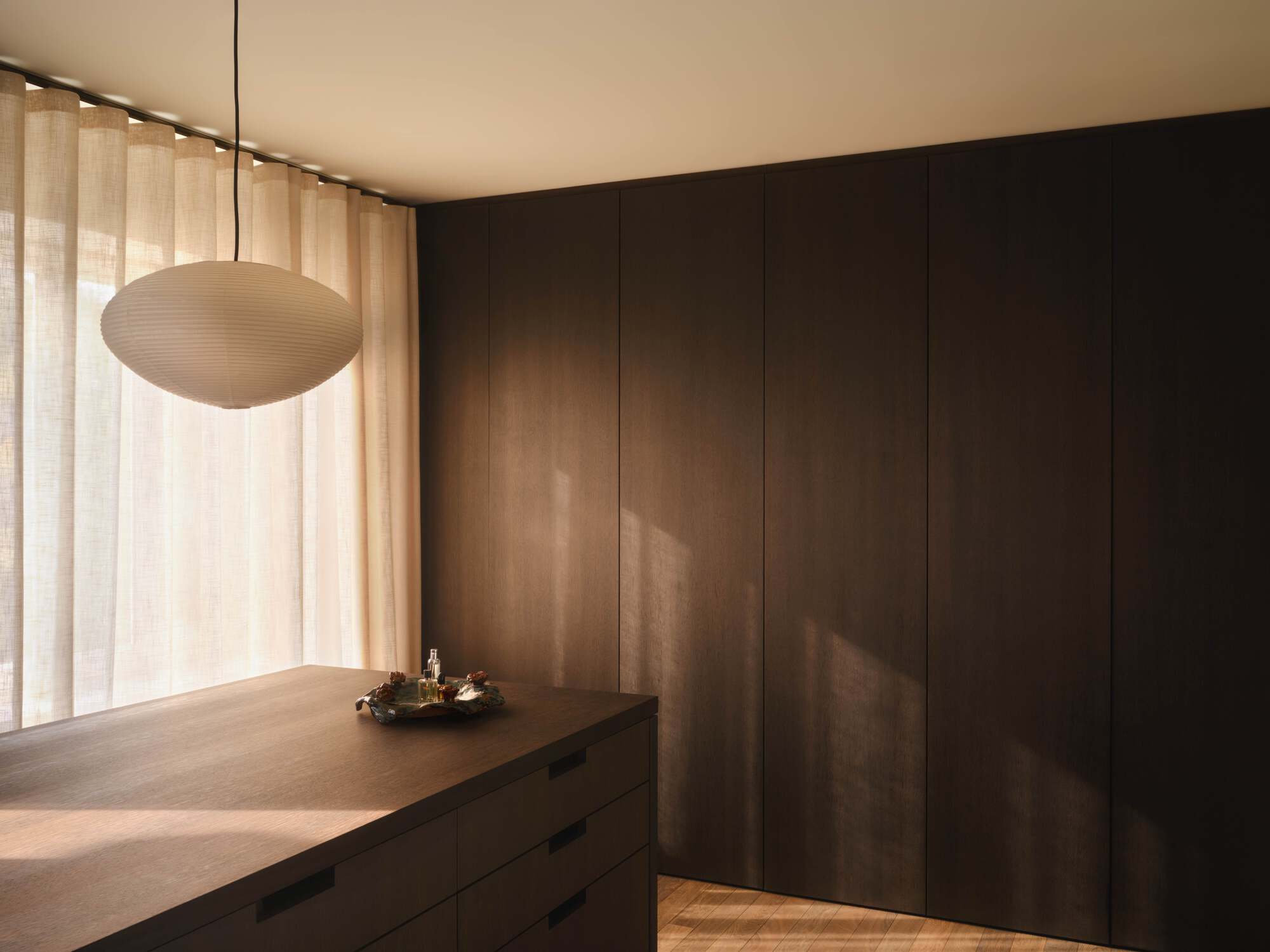 Kitchen island with oval pendant light beside sheer curtains and dark timber wall panels in afternoon sun