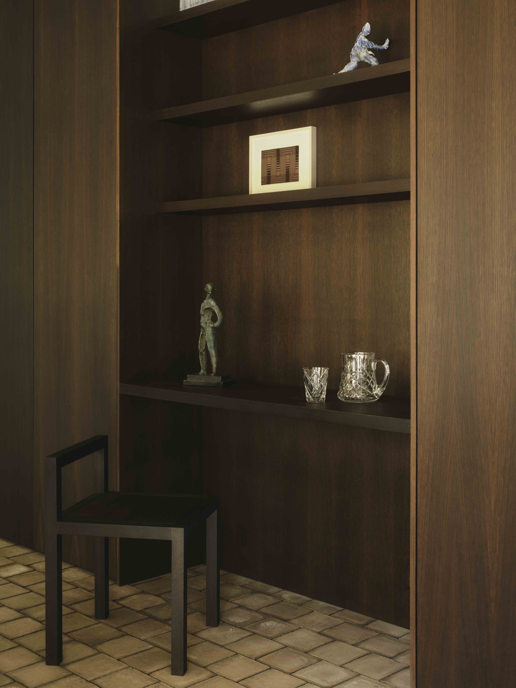 Dark timber shelving unit with display objects and small black chair against terracotta tile floor