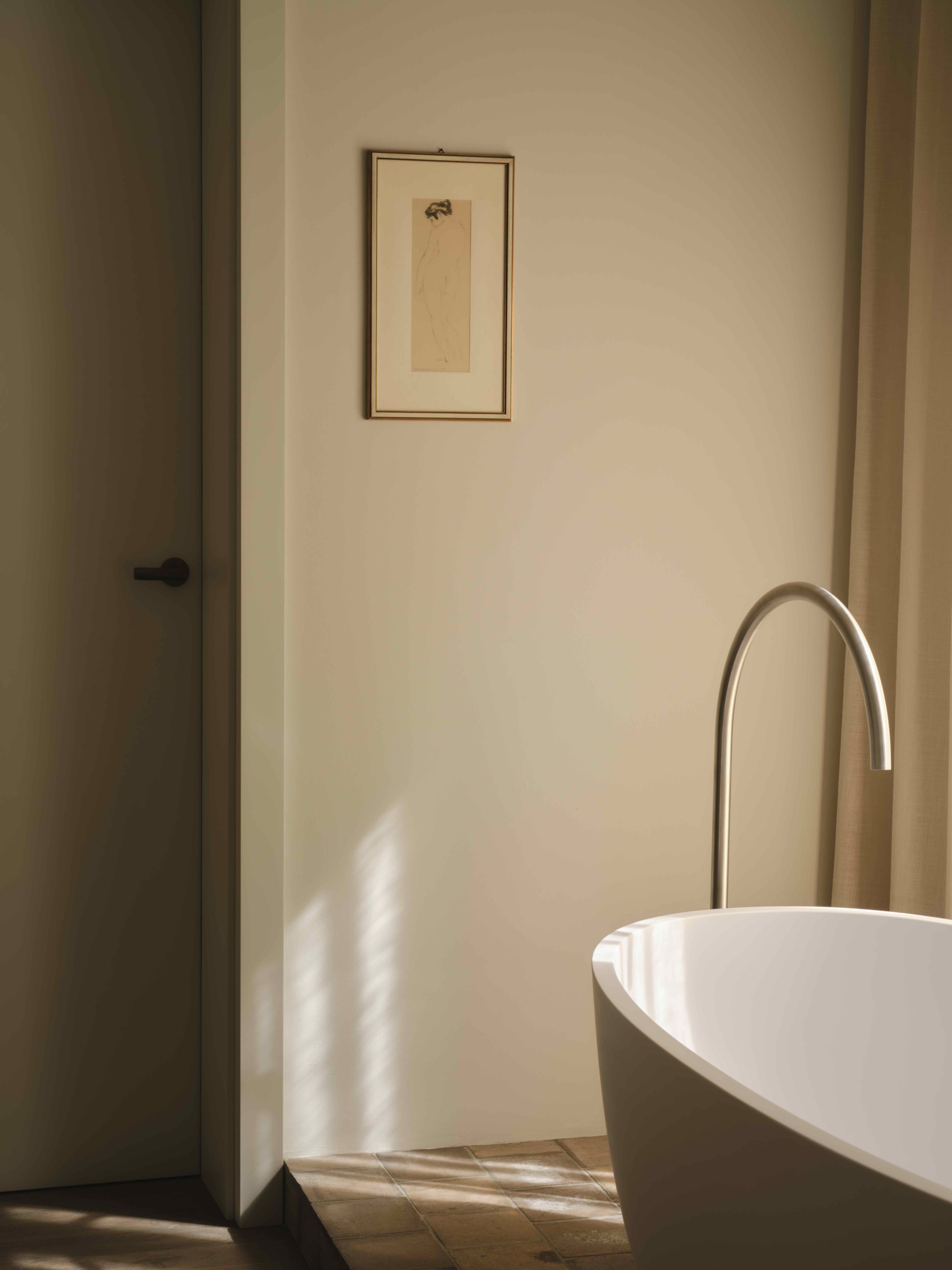 Freestanding white bathtub with arched floor-mounted faucet in soft afternoon light beside a framed drawing