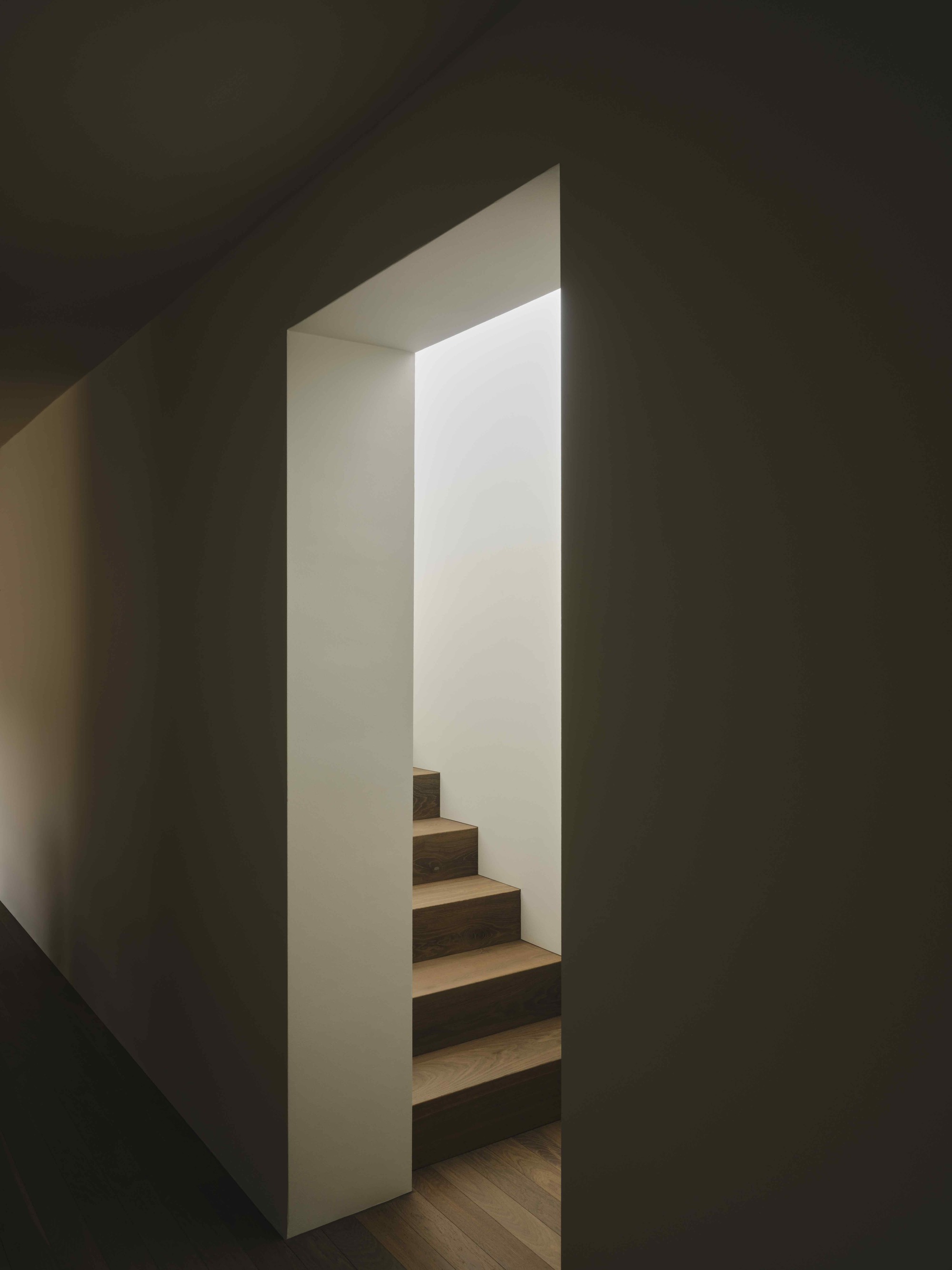 Narrow stairwell with timber treads framed by dark gray walls and natural light from above
