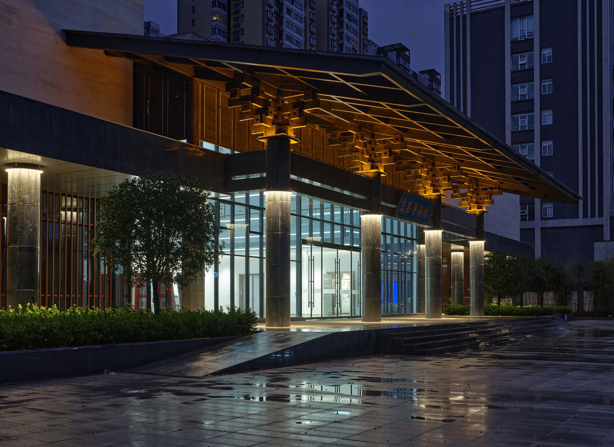 Night view of the illuminated glass entrance beneath a sculptural timber soffit canopy with wet paving reflecting light