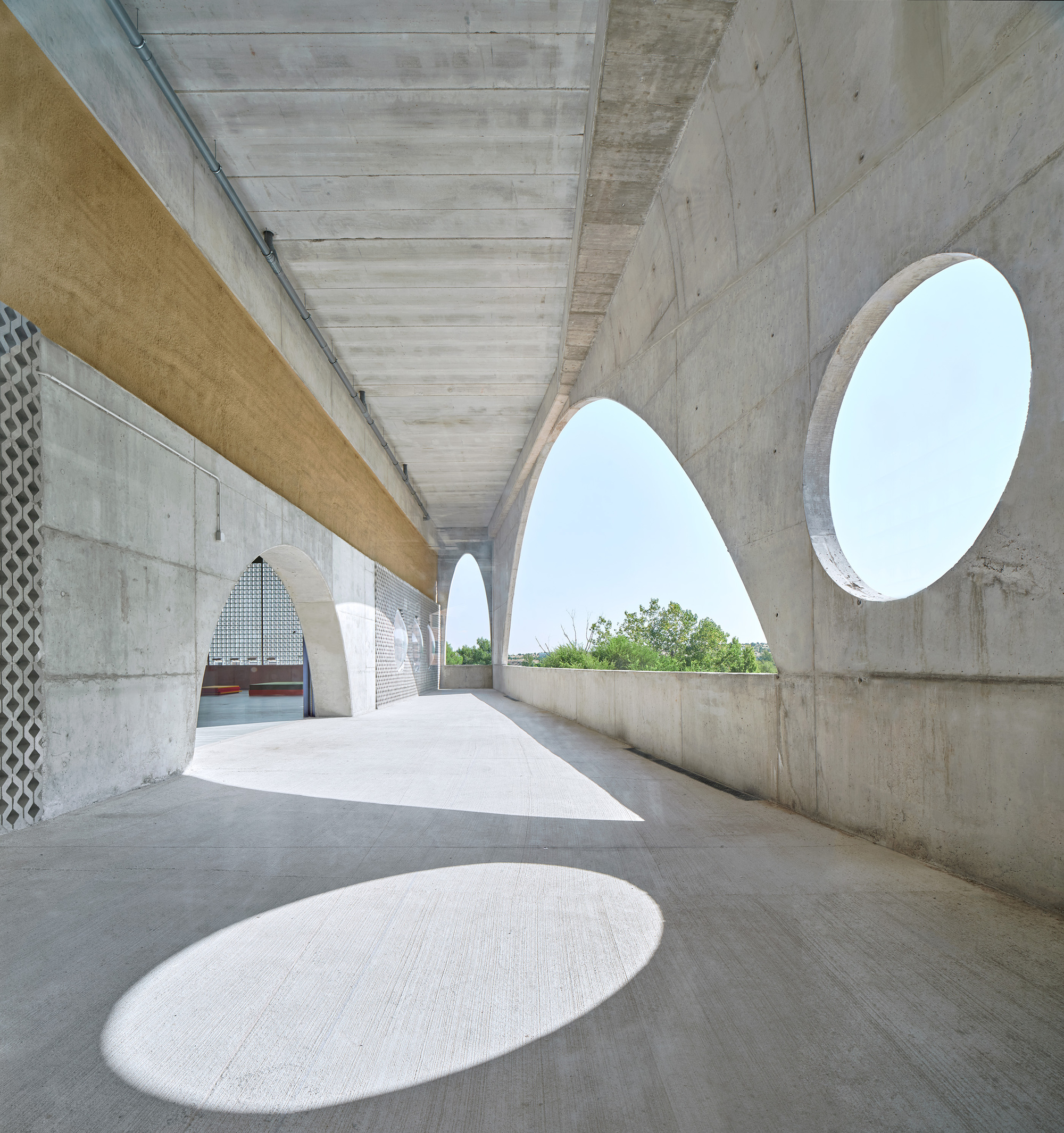 Covered outdoor corridor with boardformed concrete walls, oval openings, and sunlight casting shadows on the floor