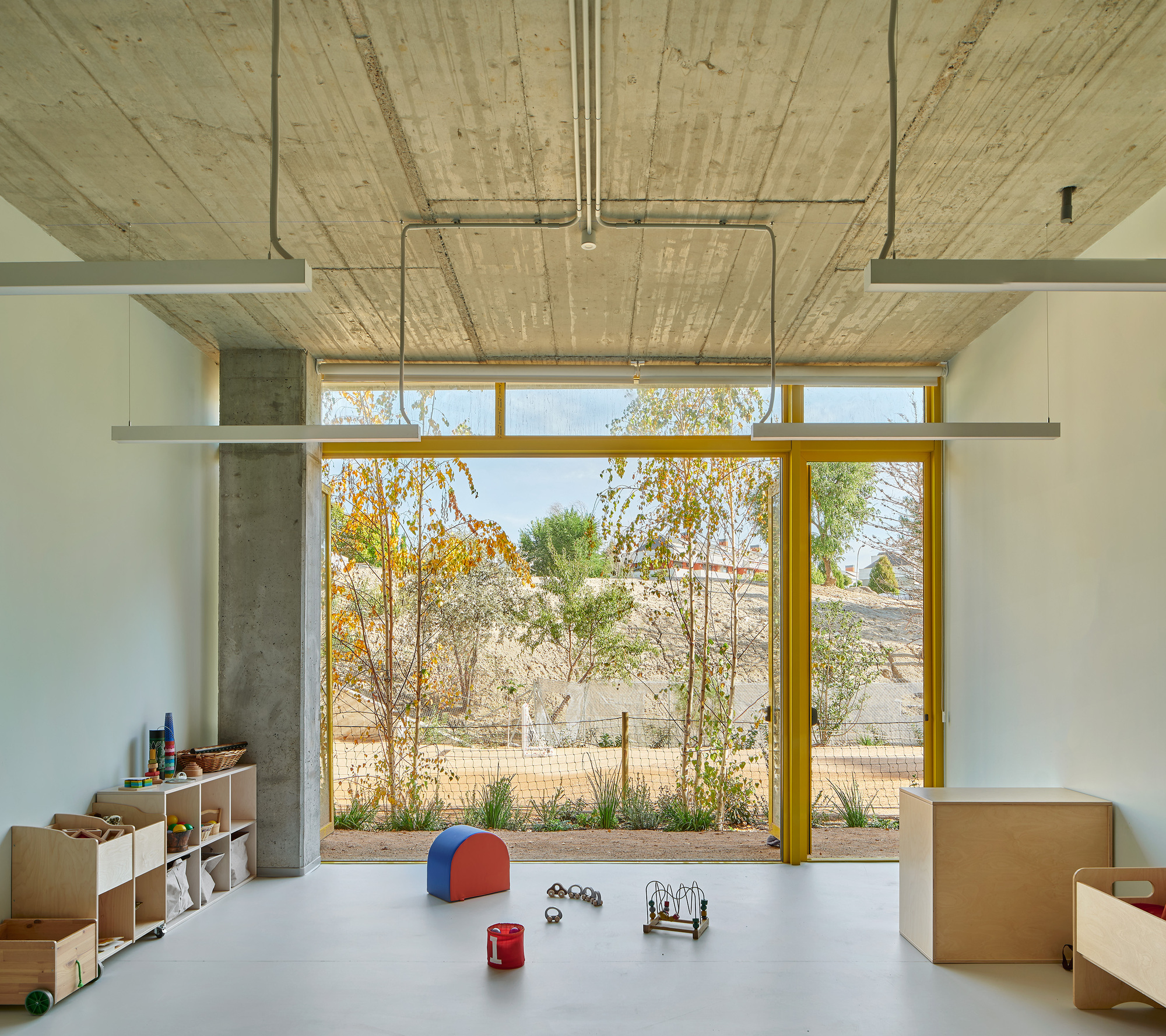 Interior classroom with yellow-framed glazing overlooking a planted courtyard and exposed concrete ceiling with conduits