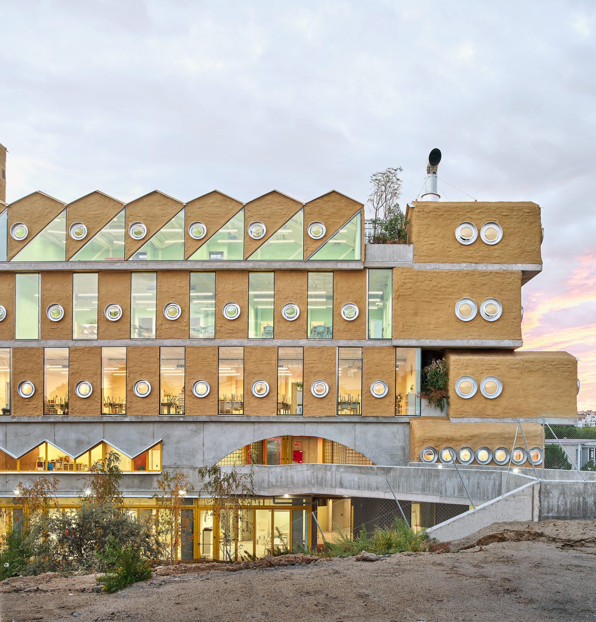 Illuminated facade at dusk showing stacked volumes with circular windows and external staircase at the base